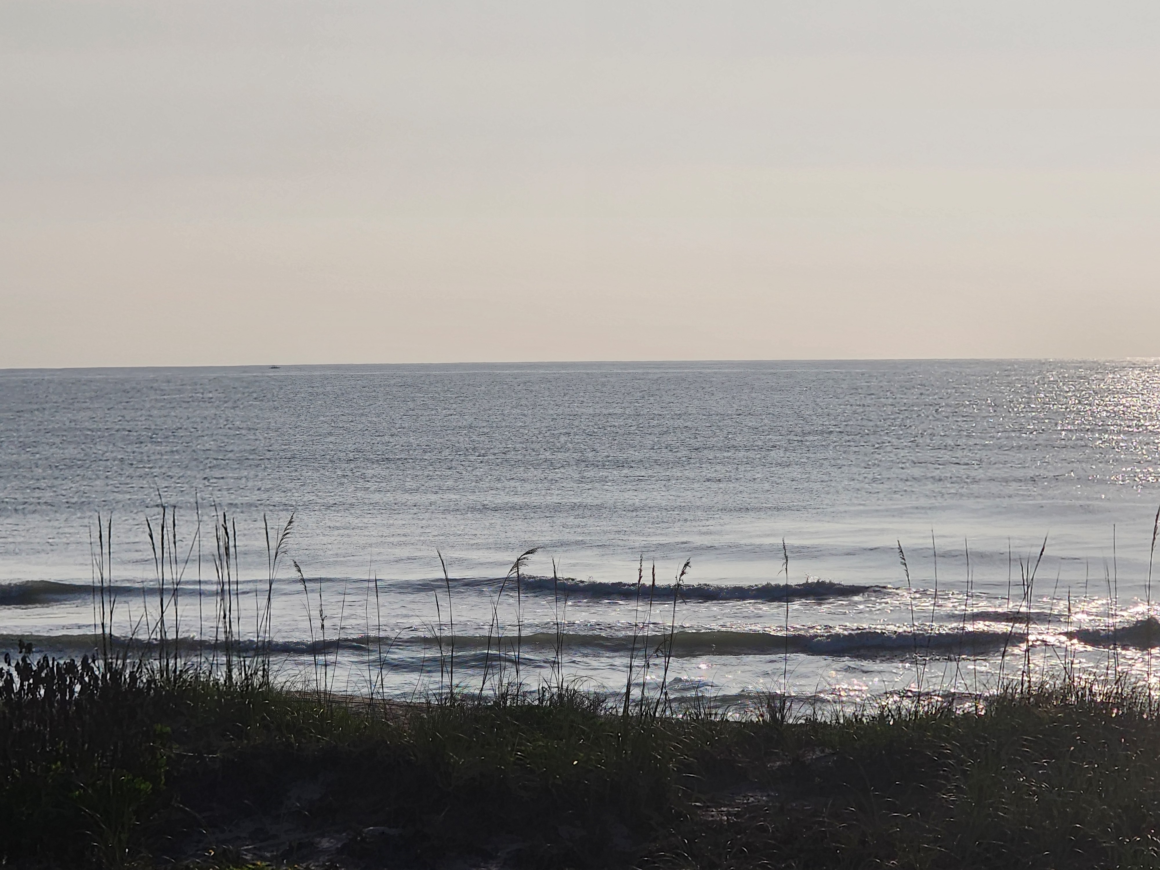 Glassy 1 foot wave breaking through silhouetted sea oats with sun sparkling off calm water at Picnic Tables South Cocoa Beach May 8 2026