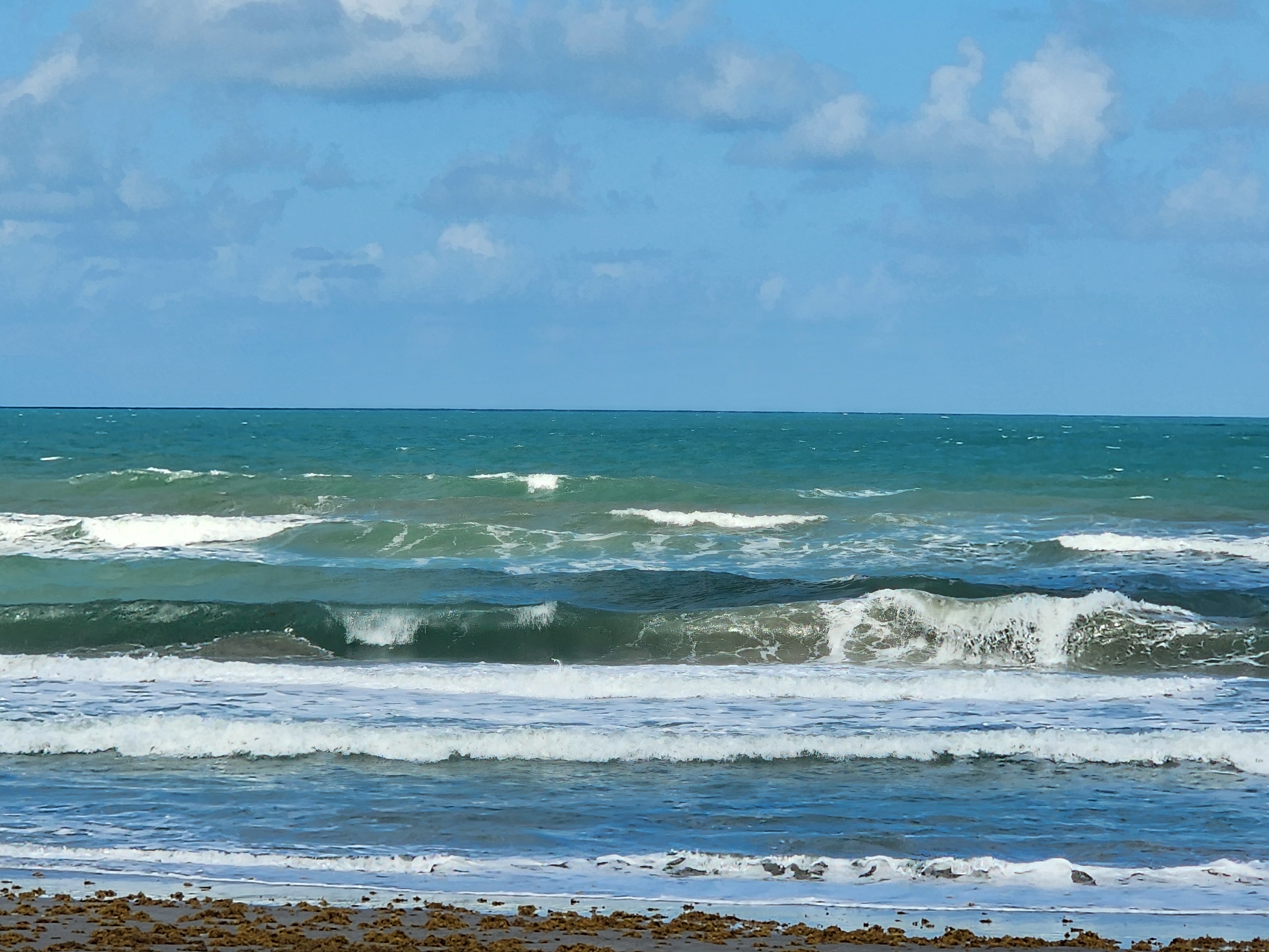 Choppy 3 foot plus afternoon surf breaking in green blue water with whitewater and sargassum along the shoreline at Picnic Tables South Cocoa Beach Tuesday May 19 2026