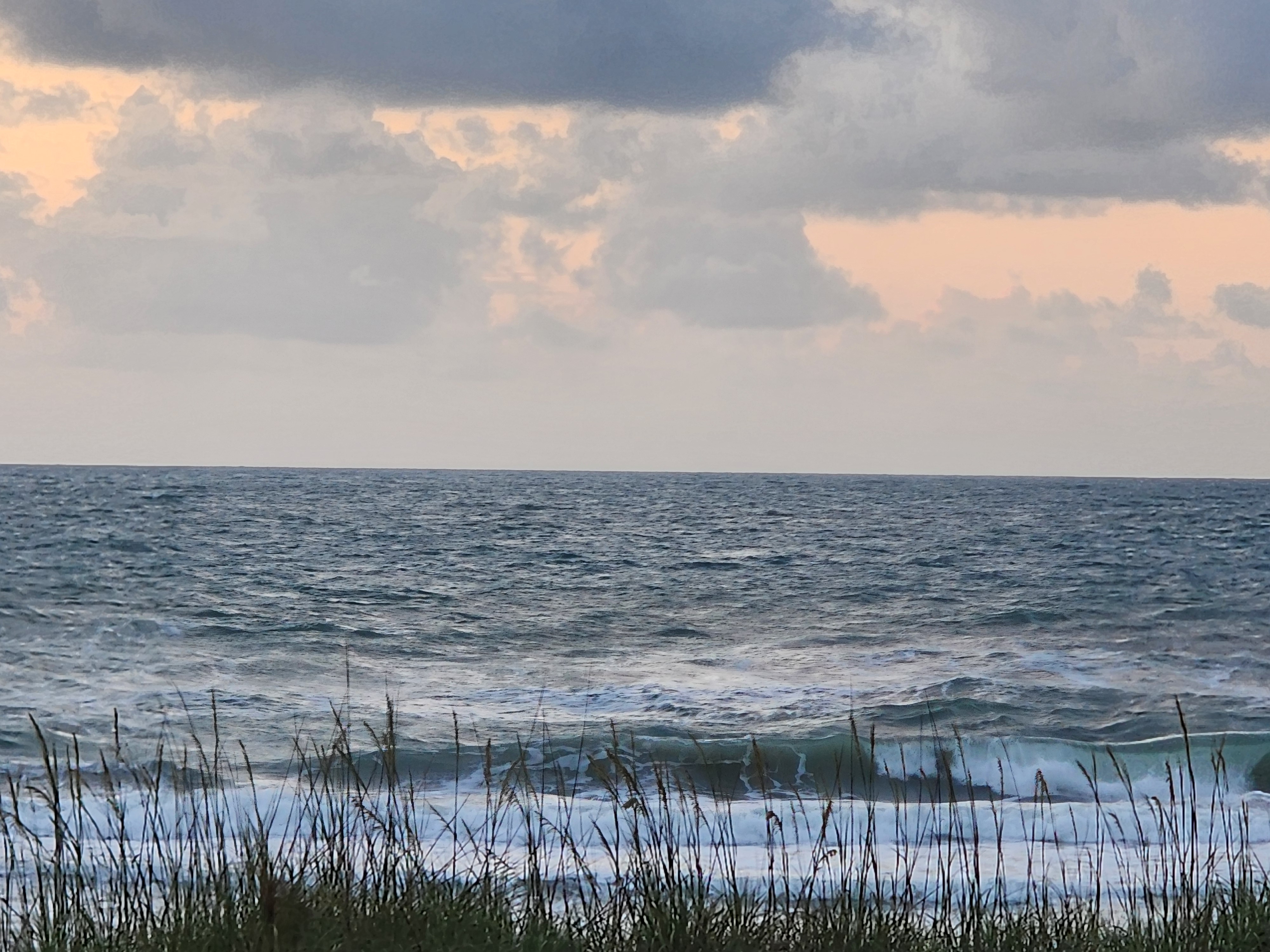 Wind-blown choppy surf with green translucent wave breaking through tall sea oats silhouetted against overcast orange sunrise at Picnic Tables South Cocoa Beach