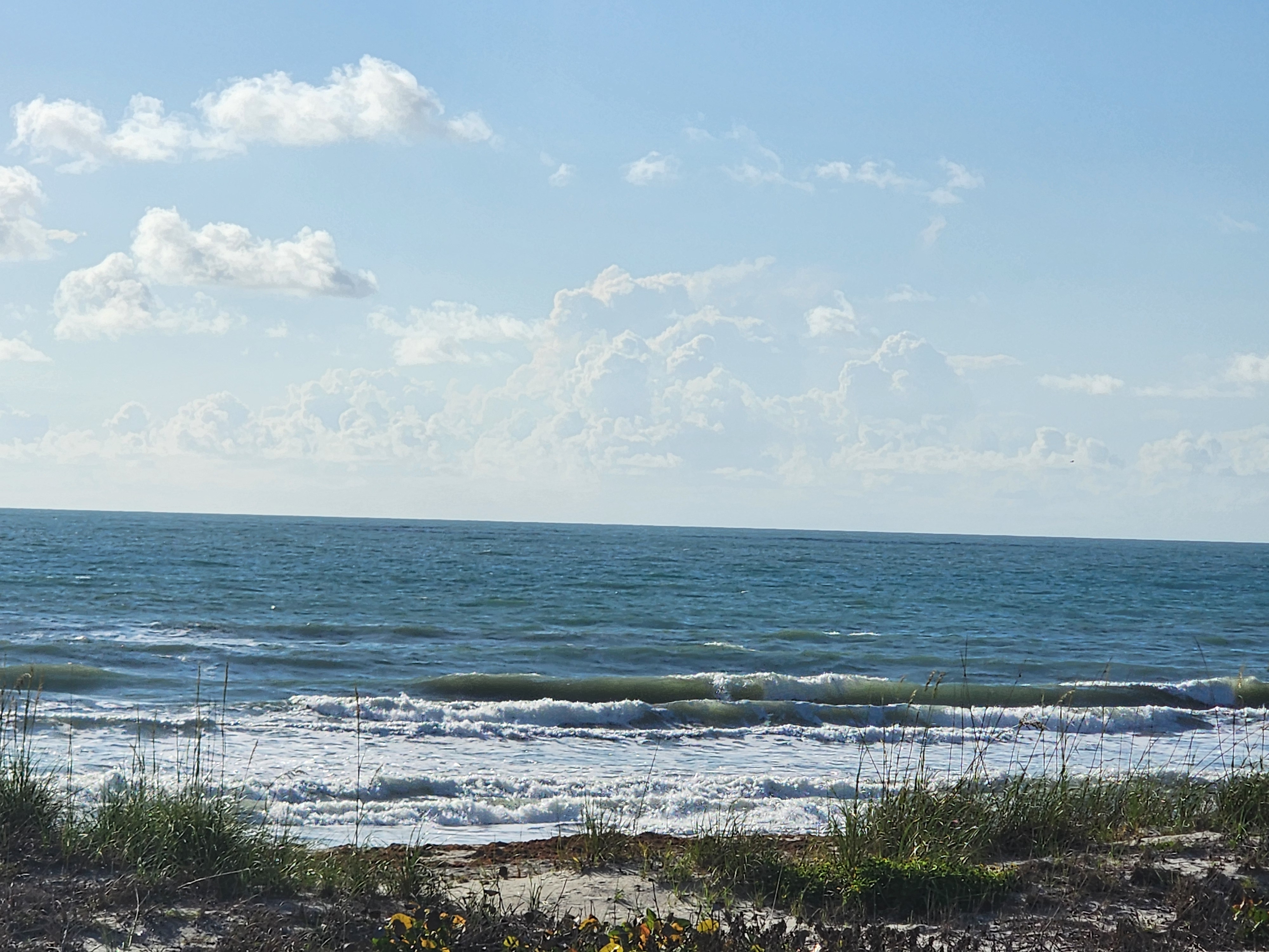 Wide view of the surf line at Cocoa Beach showing multiple waist high waves rolling in under a partly cloudy sky with sea oats along the dune line