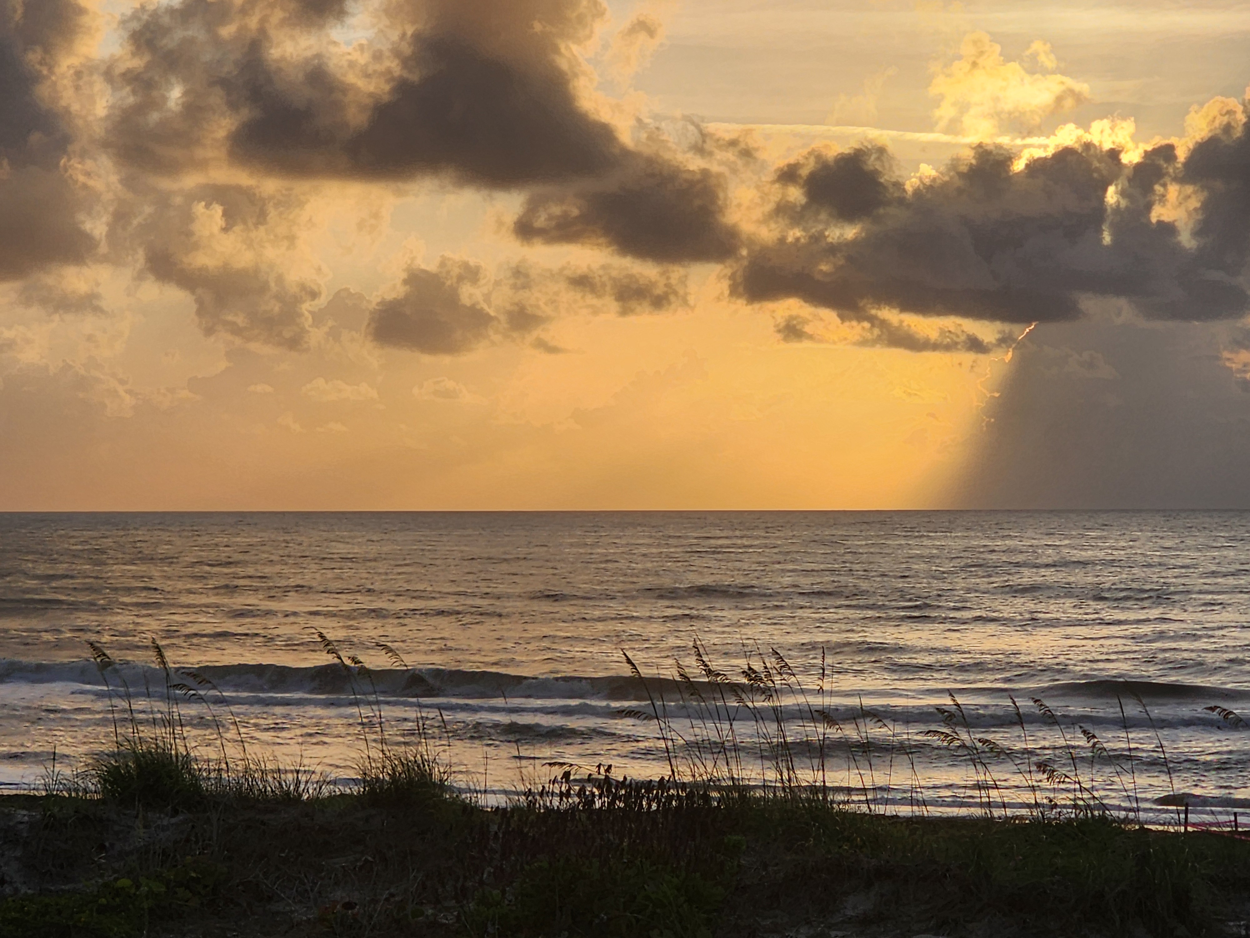 Golden sunrise with crepuscular rays breaking through dark clouds over 2-3 foot surf at South Cocoa Beach May 23 2026