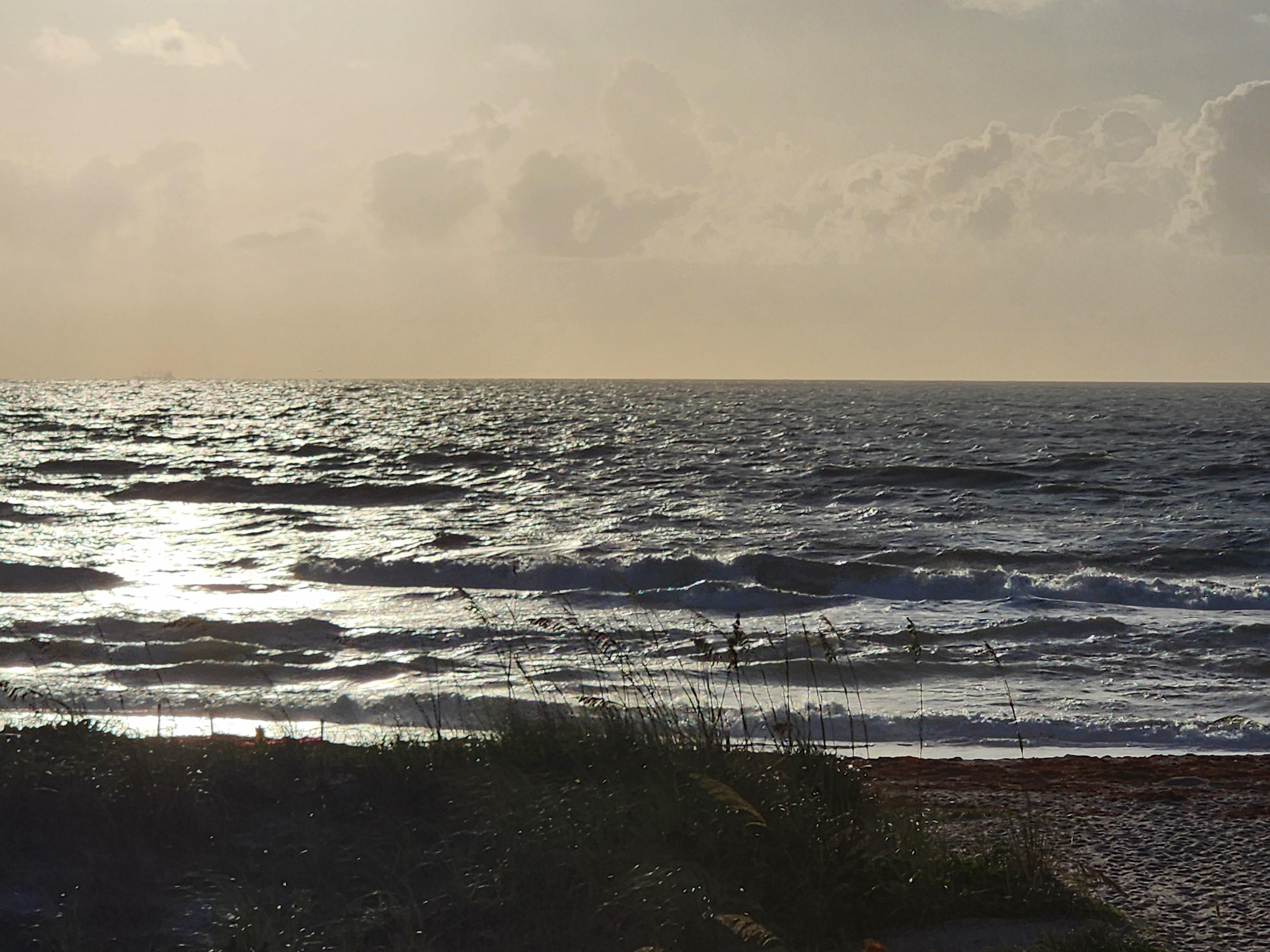 Silver sun glare over choppy wind swell with ship on horizon and silhouetted sea oats South Cocoa Beach
