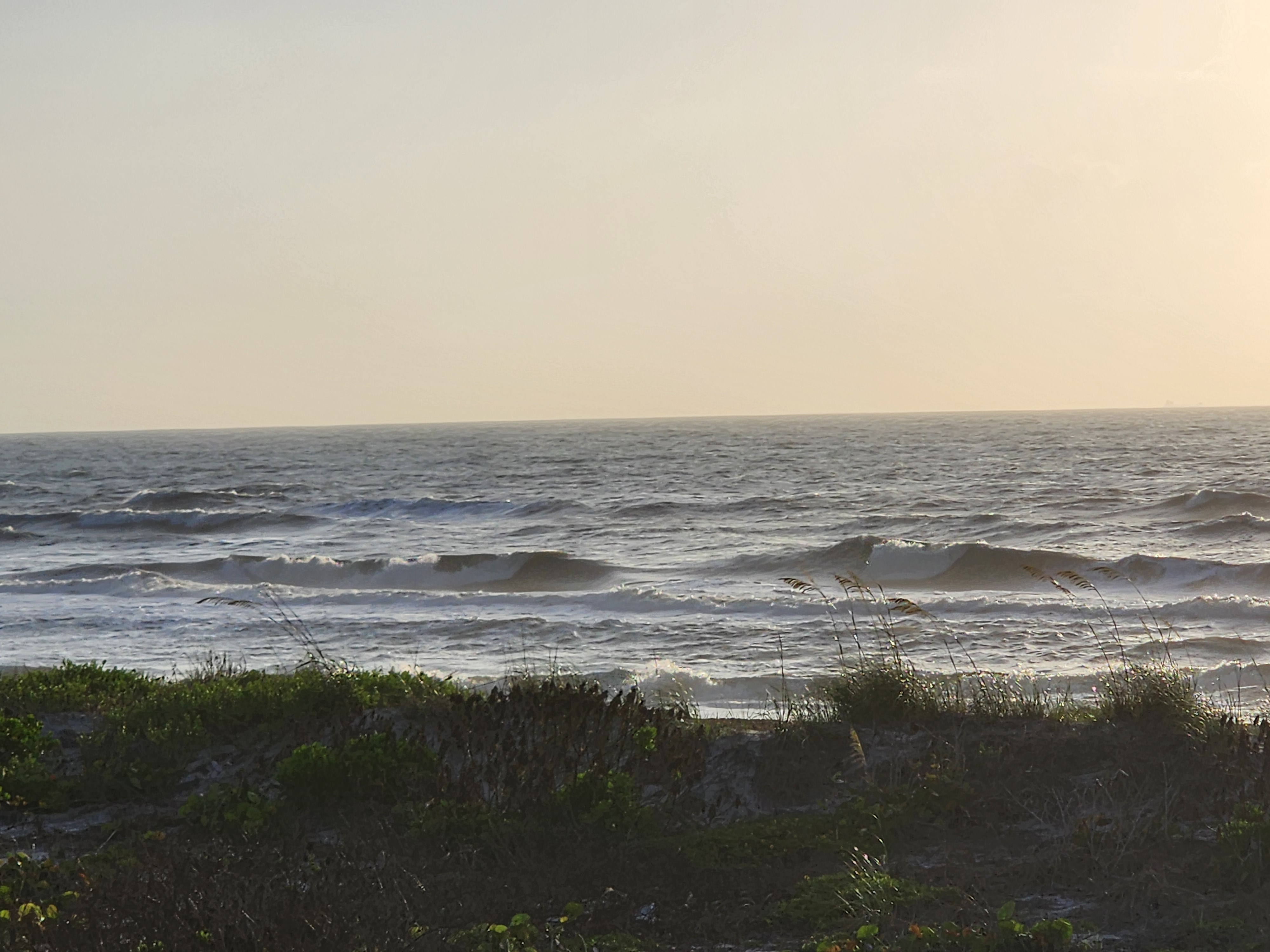 Rows of wind-textured 2-4 foot waves breaking under hazy golden skies with sea oats bending in ESE wind Cocoa Beach