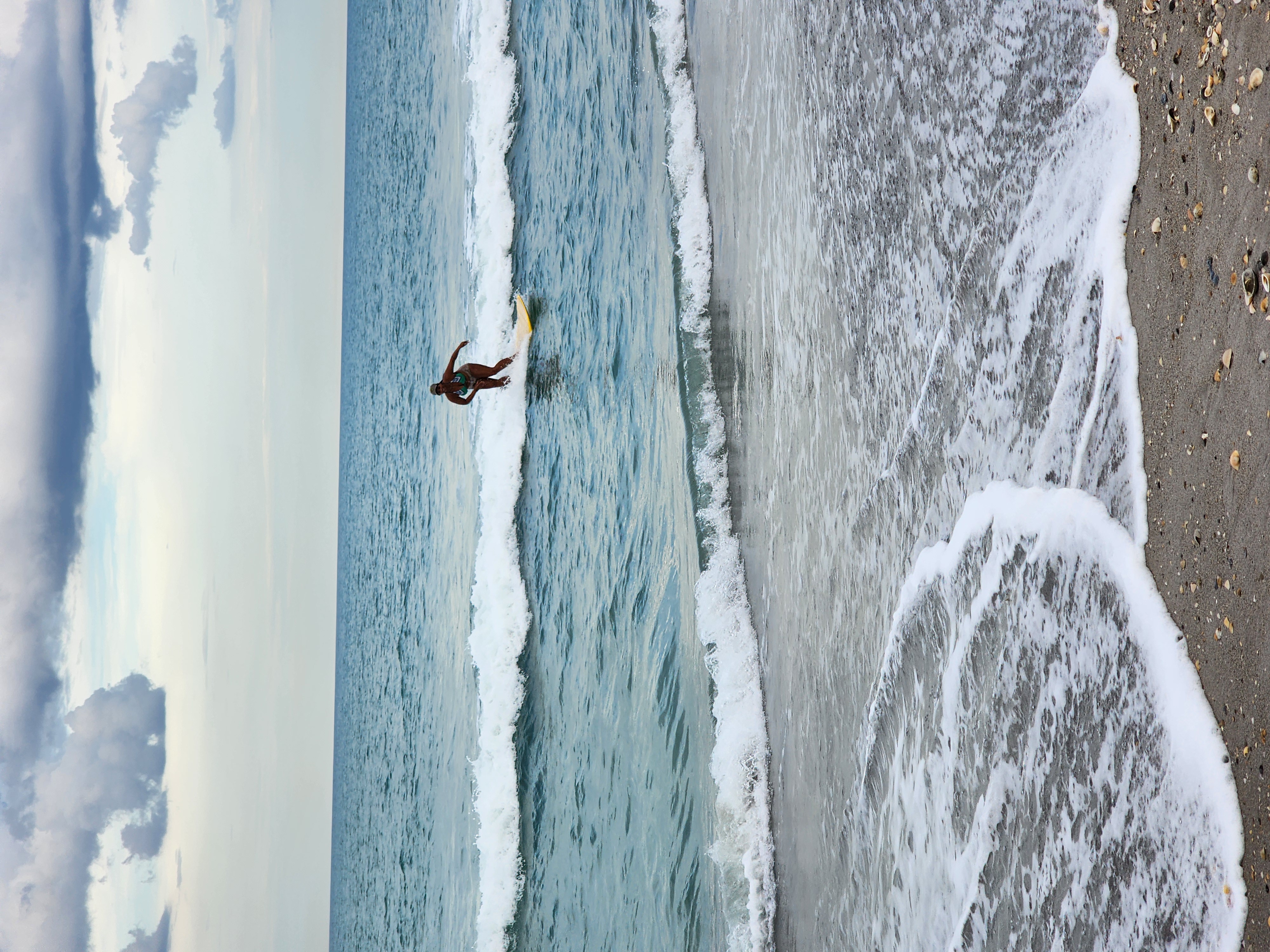 Longboarder riding a small wave in clear turquoise water with shells on the beach at Picnic Tables Cocoa Beach