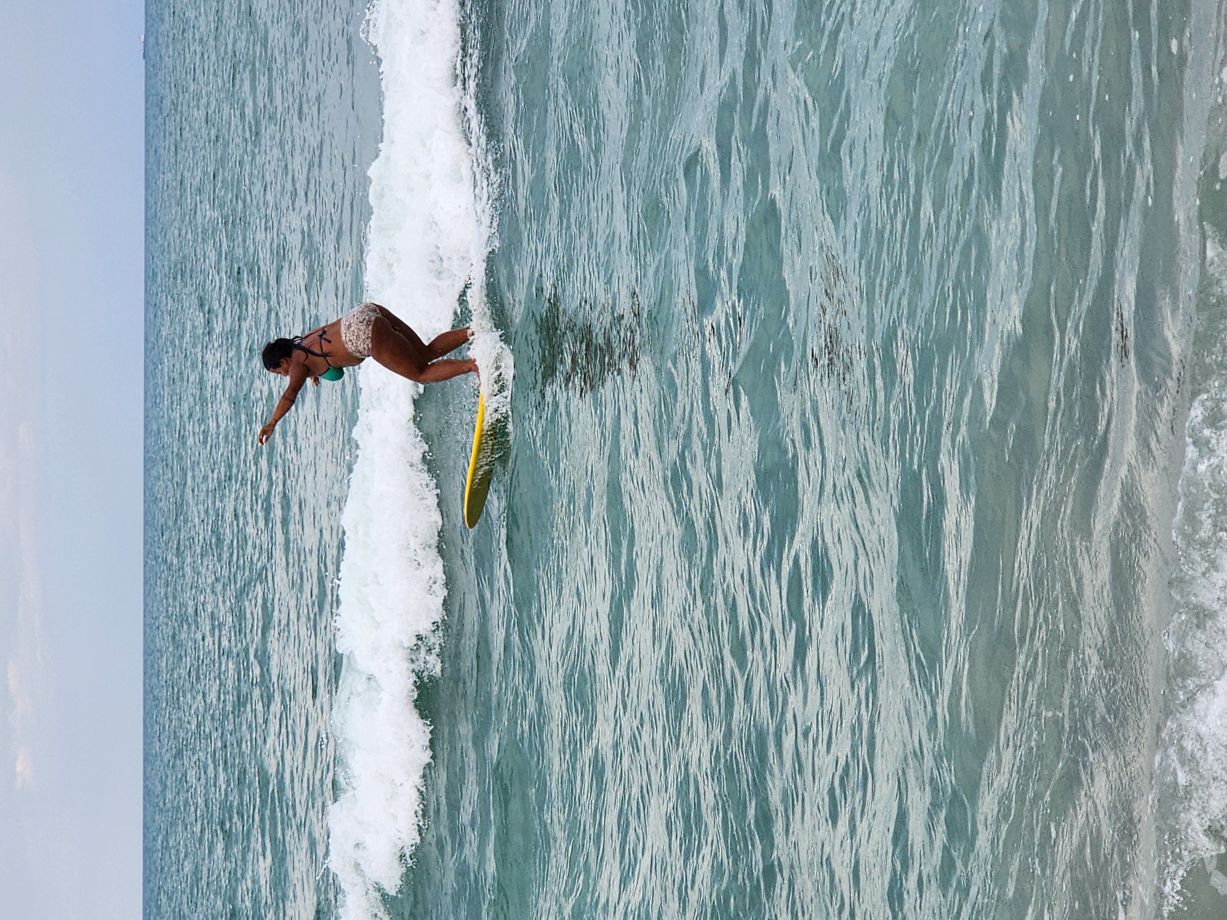 Surfer styling on yellow longboard with arm extended riding a small wave in turquoise water at Picnic Tables Cocoa Beach