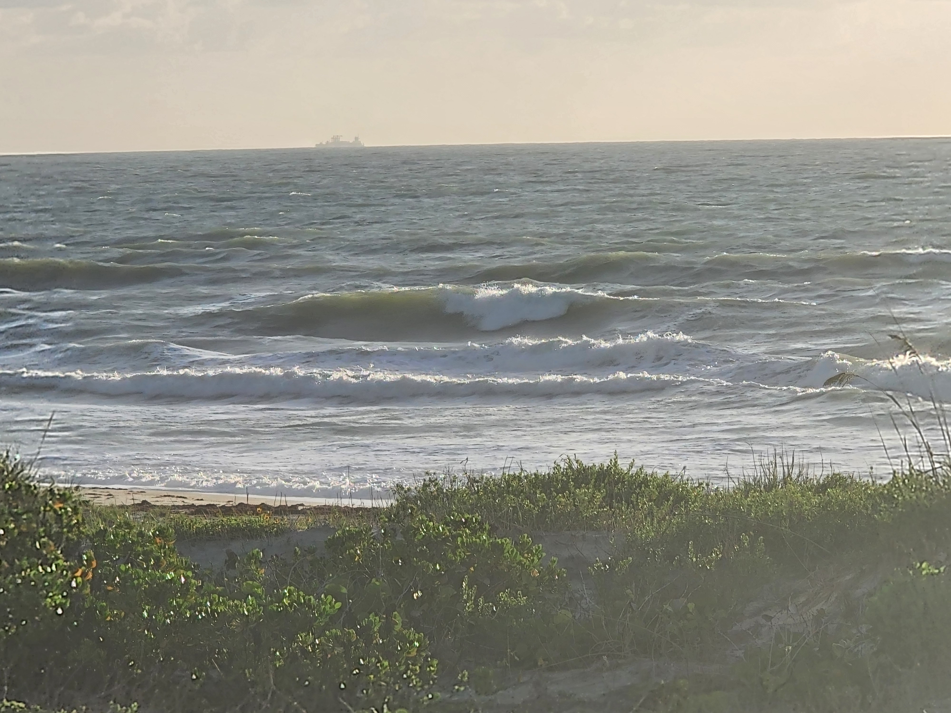 Solid green wave face breaking through choppy conditions with ship on horizon and dune vegetation at Picnic Tables Cocoa Beach