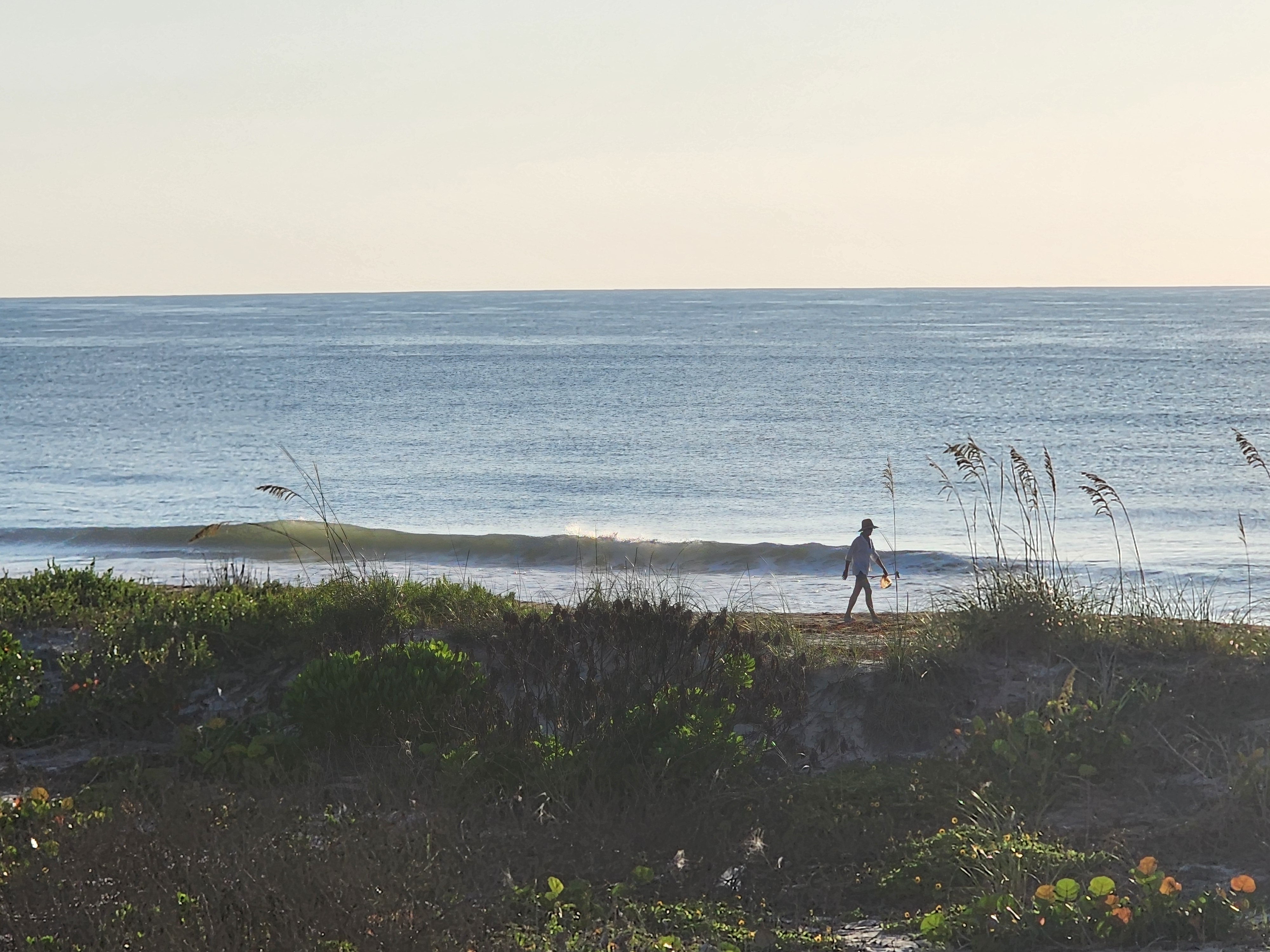 Beachgoer walking the shoreline past a tiny clean wave at South Cocoa Beach, light west wind morning