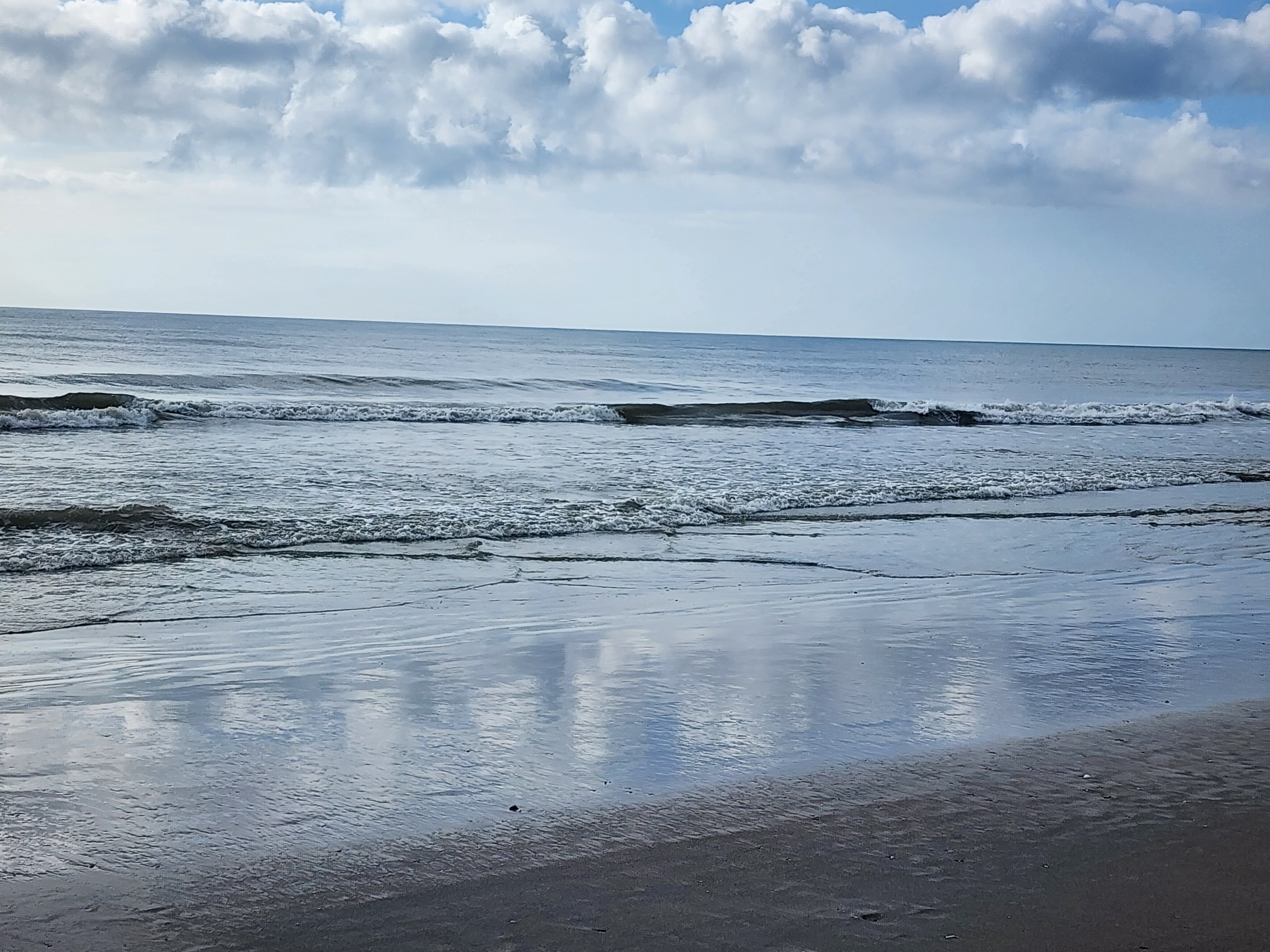 Wide low-tide view with puffy cumulus clouds reflected in the wet sand and clean wave lines breaking across the sandbar