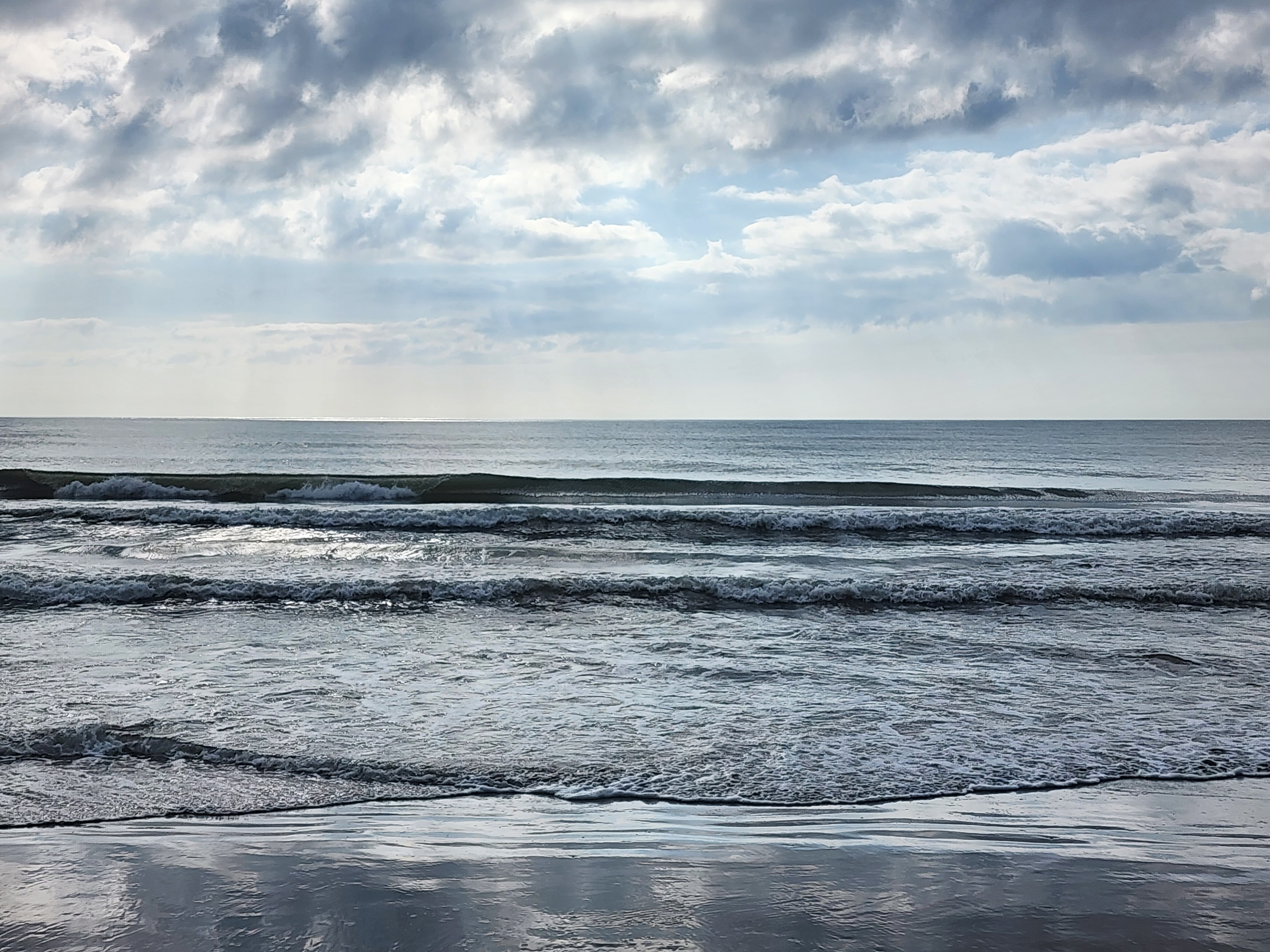 Several layered lines of small clean waves rolling in over a wide low-tide flat, cloudy sky reflected on the glassy sand