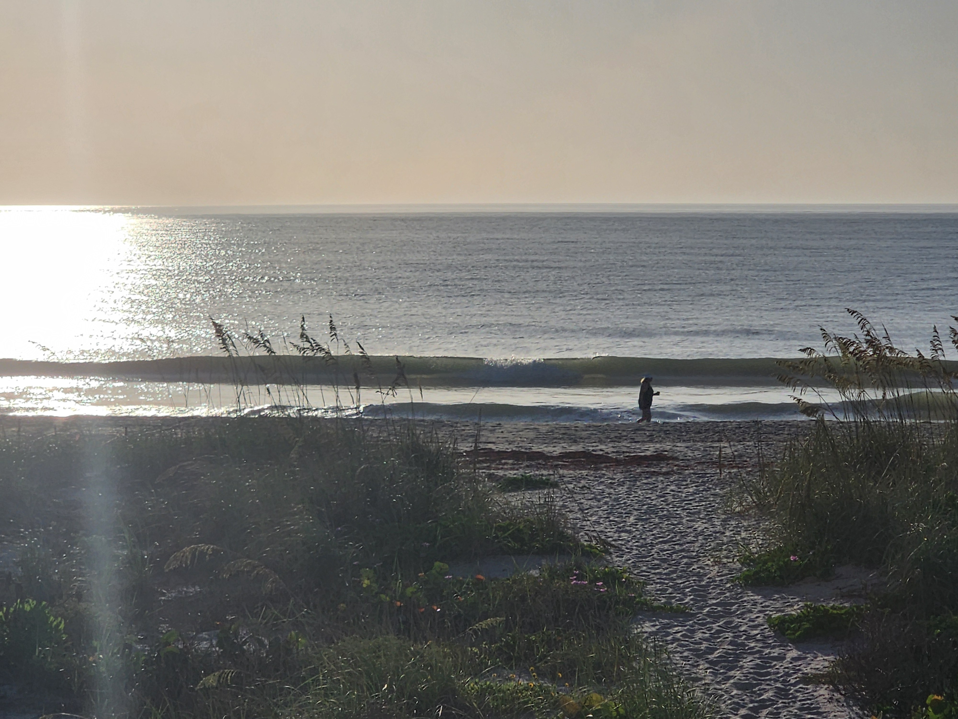 Backlit morning view down the dune path to the beach, sun glinting silver off the water and a lone beachcomber walking the shoreline as a small wave breaks behind them.