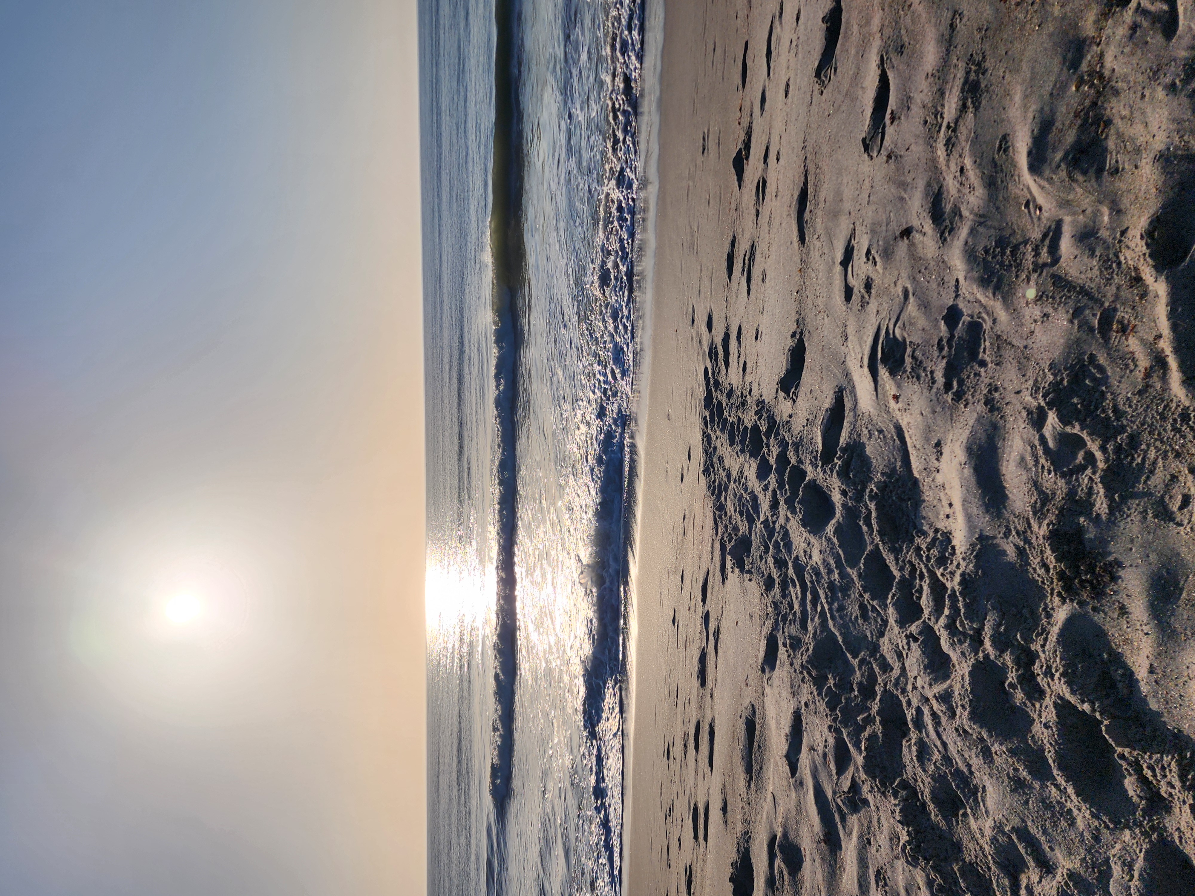 Low morning sun throwing a bright glare path across the water, a clean wave breaking and turtle-crawl tracks pressed into the wet sand in the foreground.