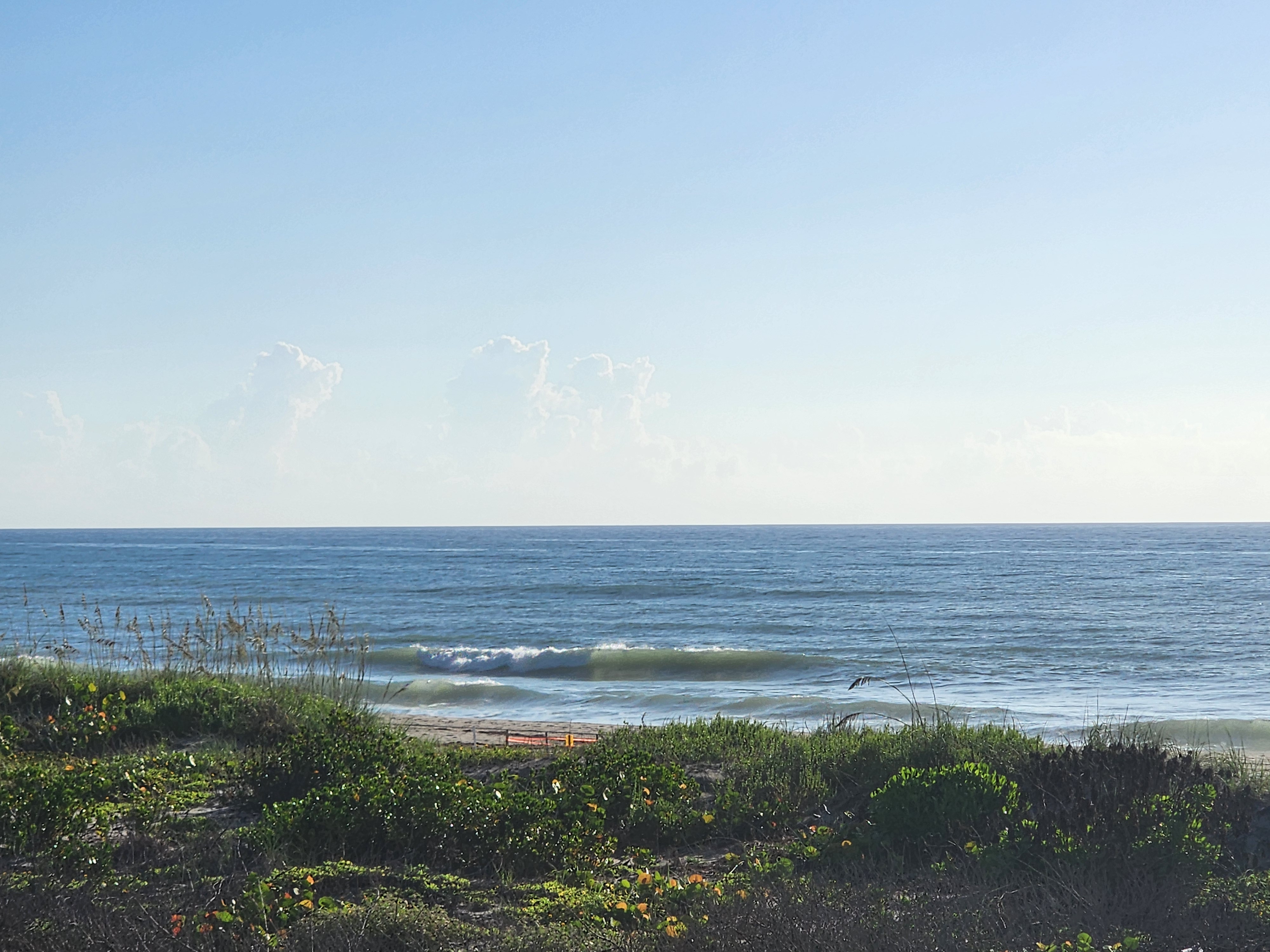 Wide view of the Atlantic on the incoming tide, small set wave breaking close to shore under a bright blue sky with distant cloud towers