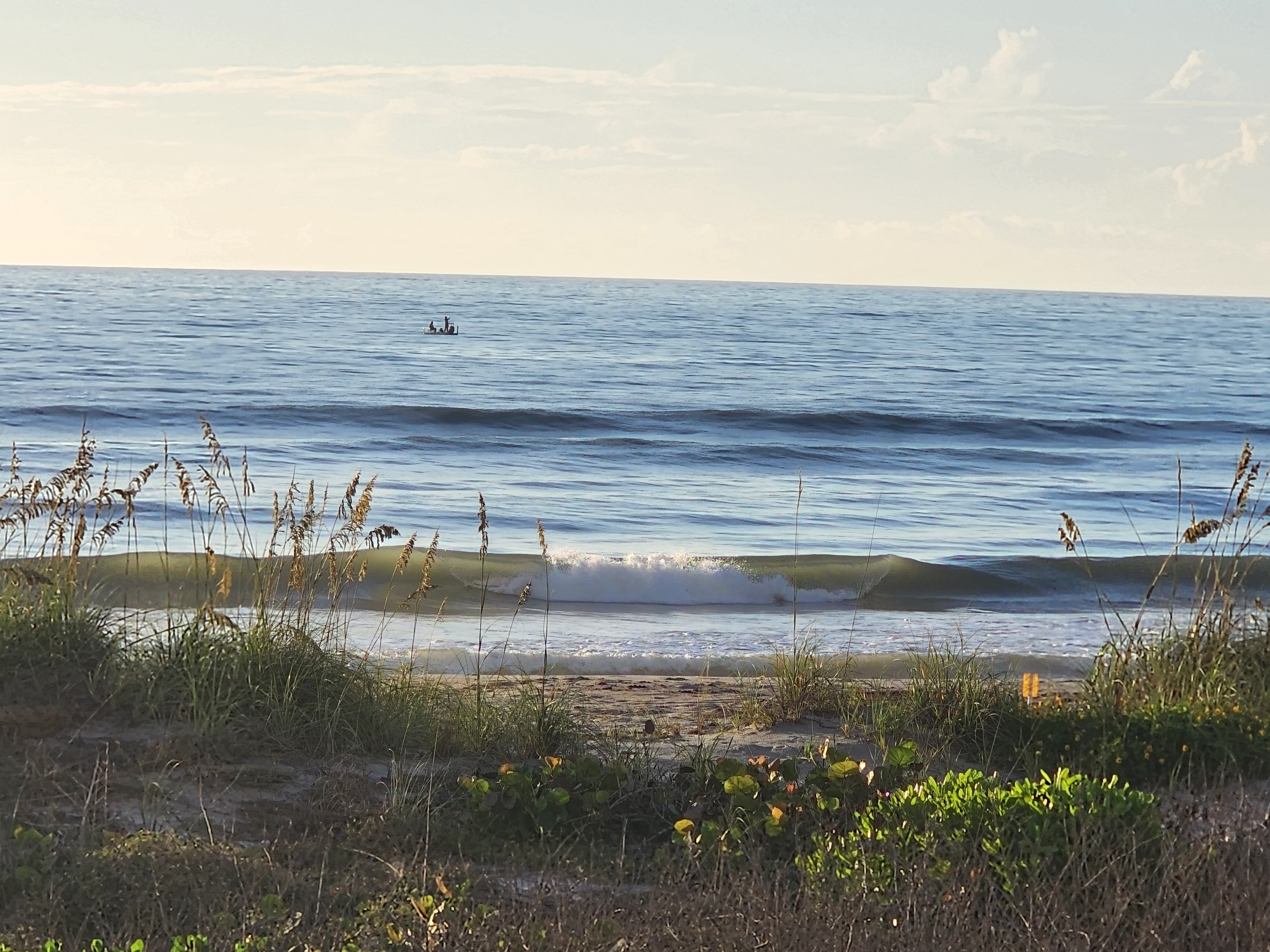 A clean chest-high wave peeling and breaking at Picnic Tables, framed by golden sea oats in the foreground with a small skiff of anglers anchored out past the lineup.