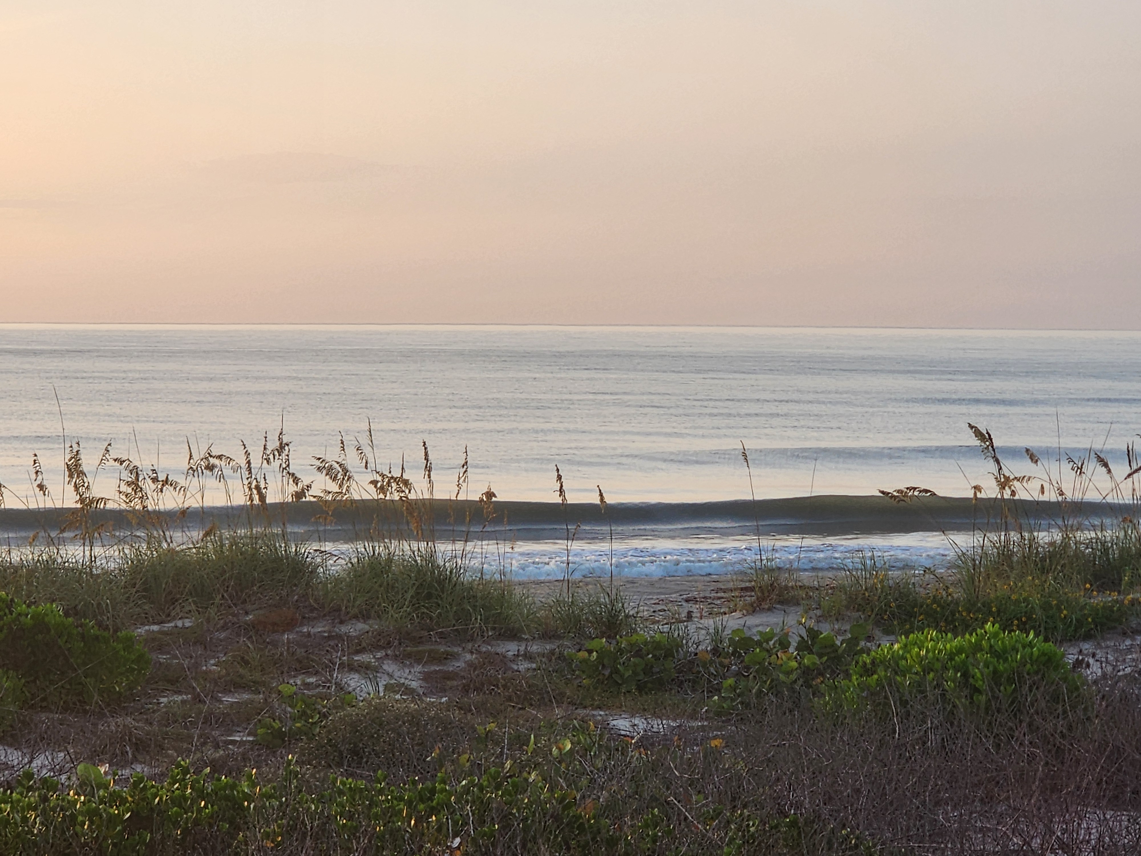 Small wave closing out near shore under a hazy pink sunrise sky, South Cocoa Beach, July 2026
