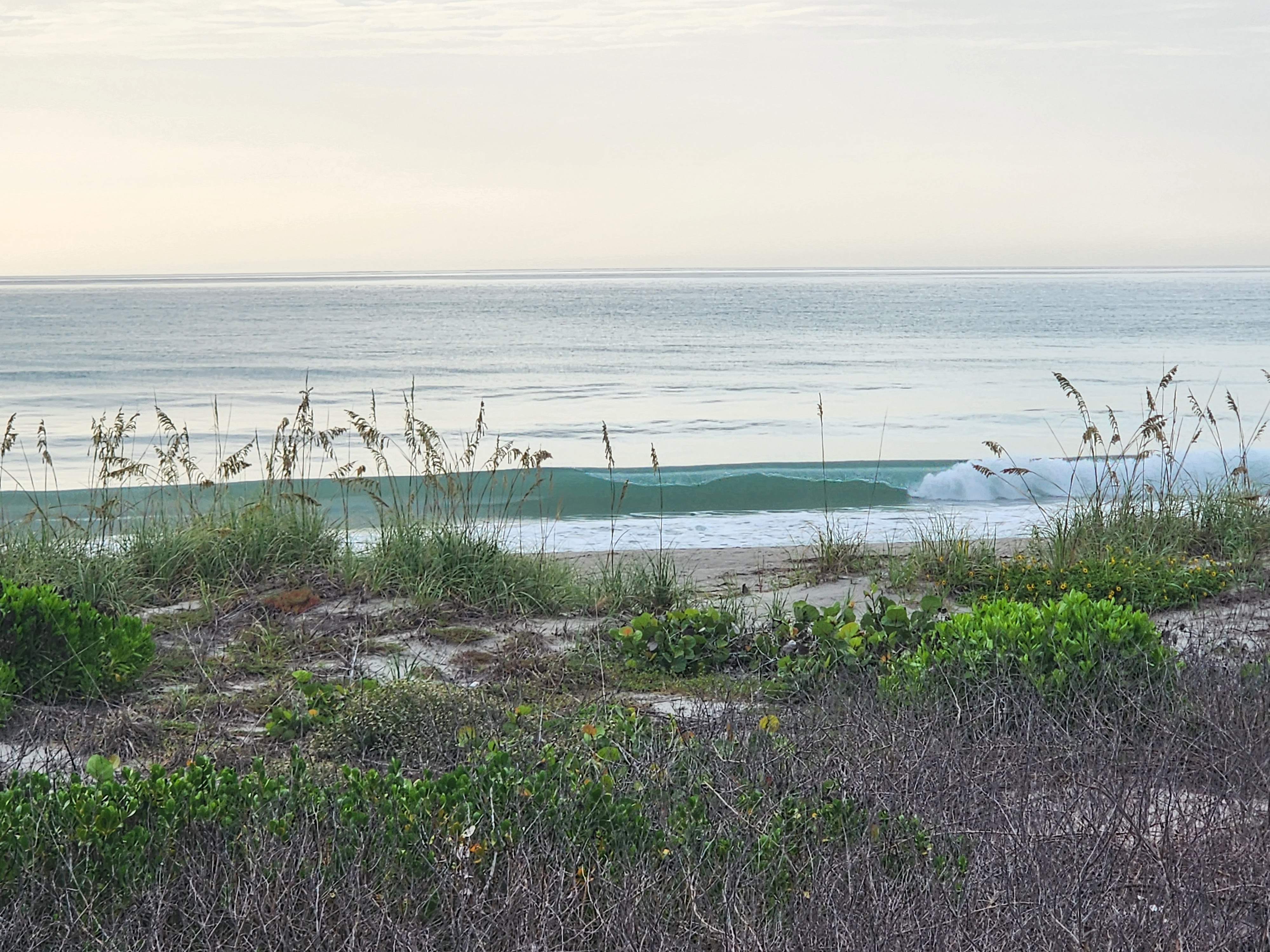 Overcast sky over calm water with a small wave breaking near the shoreline, South Cocoa Beach, July 2026
