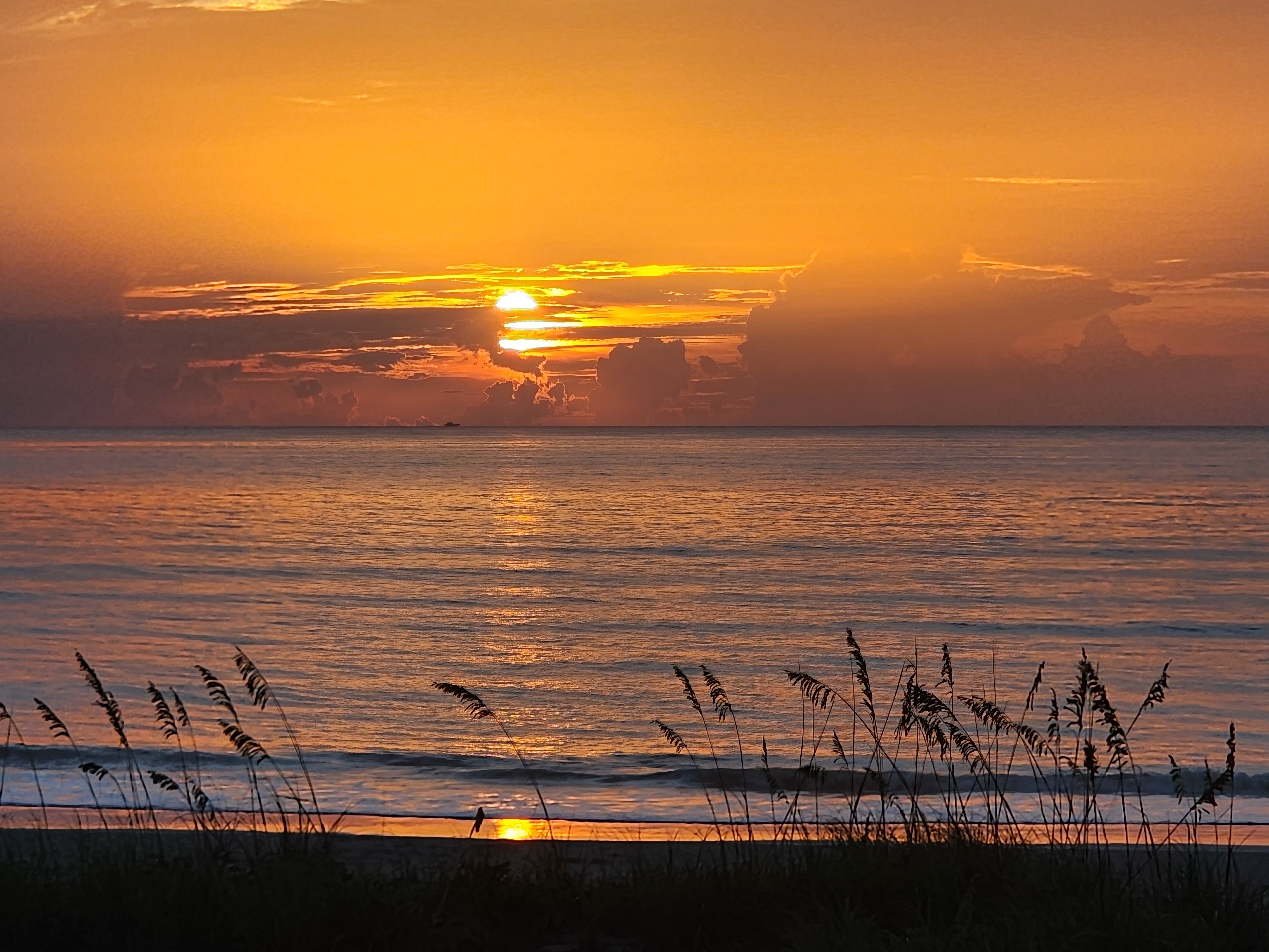 Sun breaking through a low cloud bank at sunrise, glowing orange over the water with sea oats silhouetted in the foreground