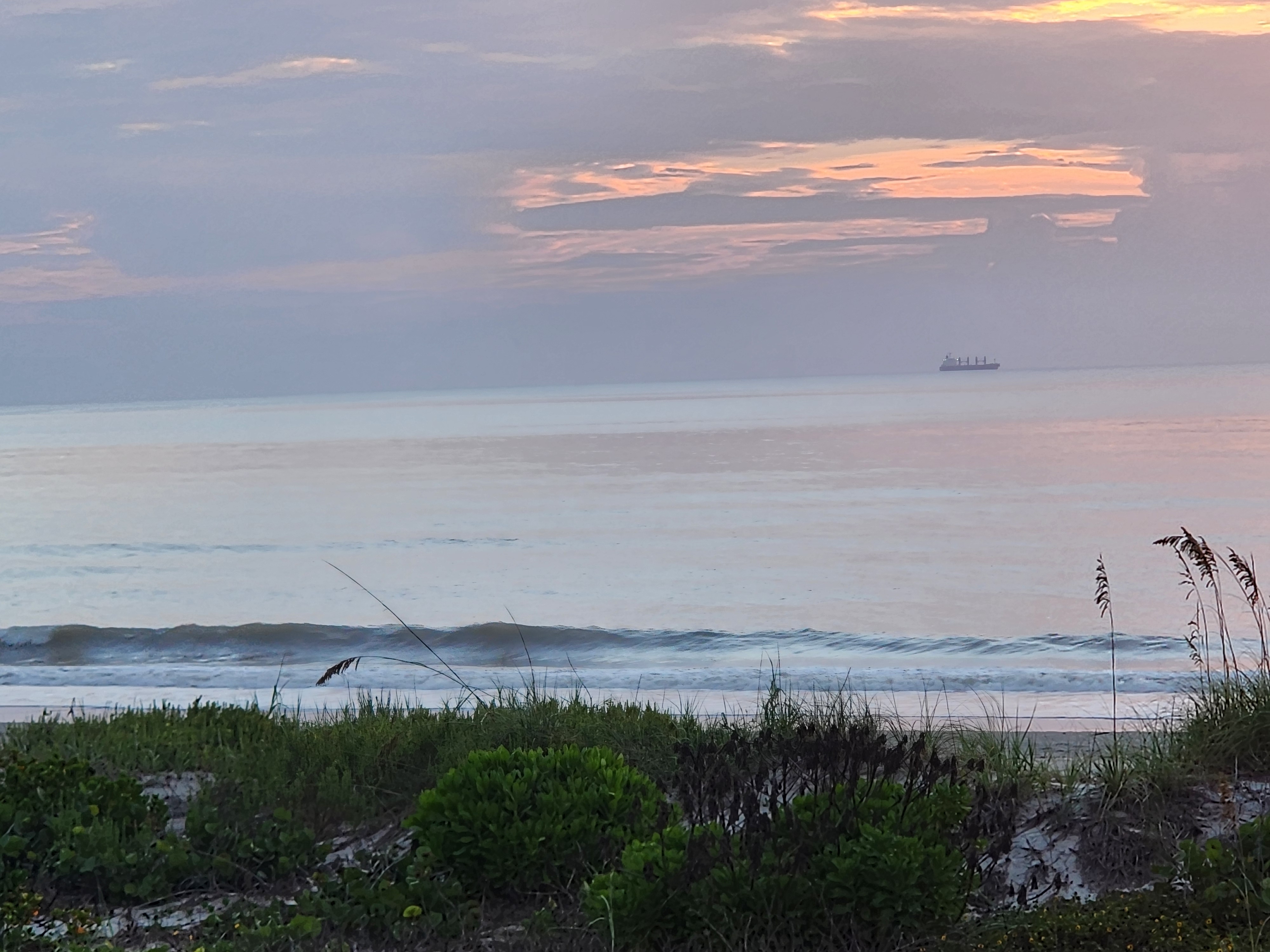 Soft pink and lavender post sunrise light over the water with a cargo ship on the horizon and a small line breaking on the sandbar