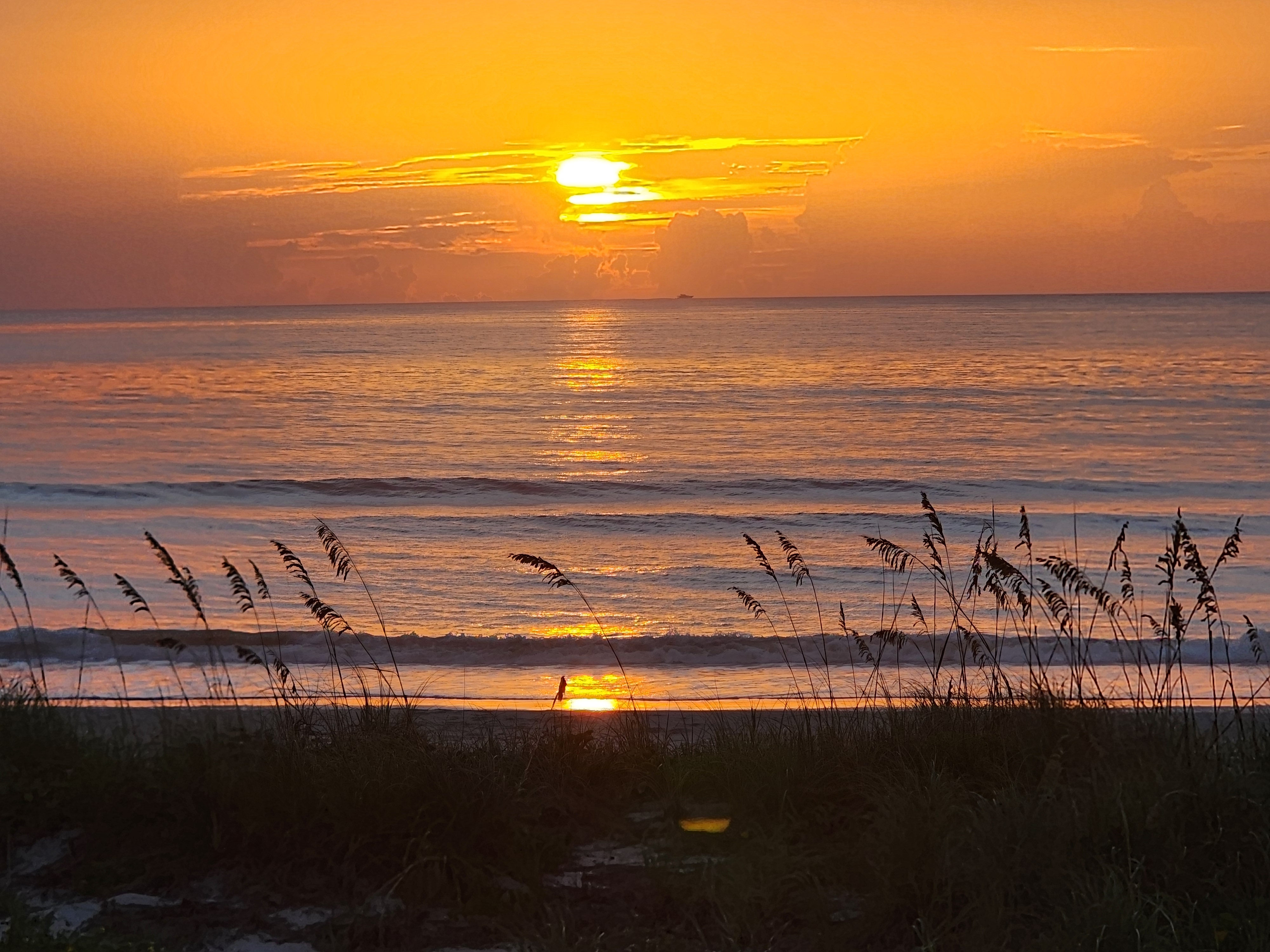 Full sun above the cloud bank casting a long gold reflection across the water, a lone beachgoer silhouetted at the waterline