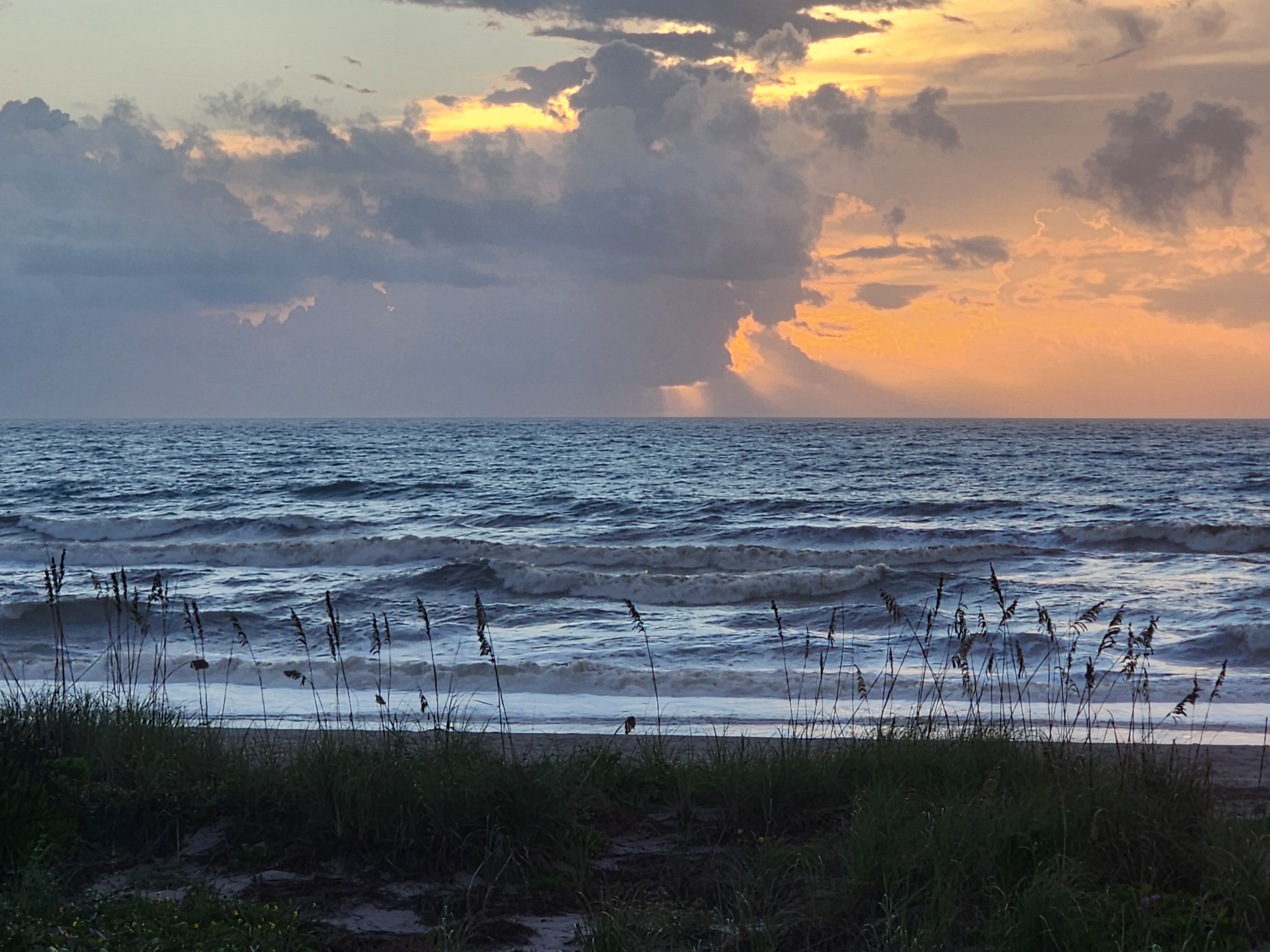 Sunrise over the Atlantic at 32nd Street with a towering storm cloud lit orange and gold, rain shafts falling on the horizon and lines of 2-4 foot surf rolling in behind silhouetted sea oats