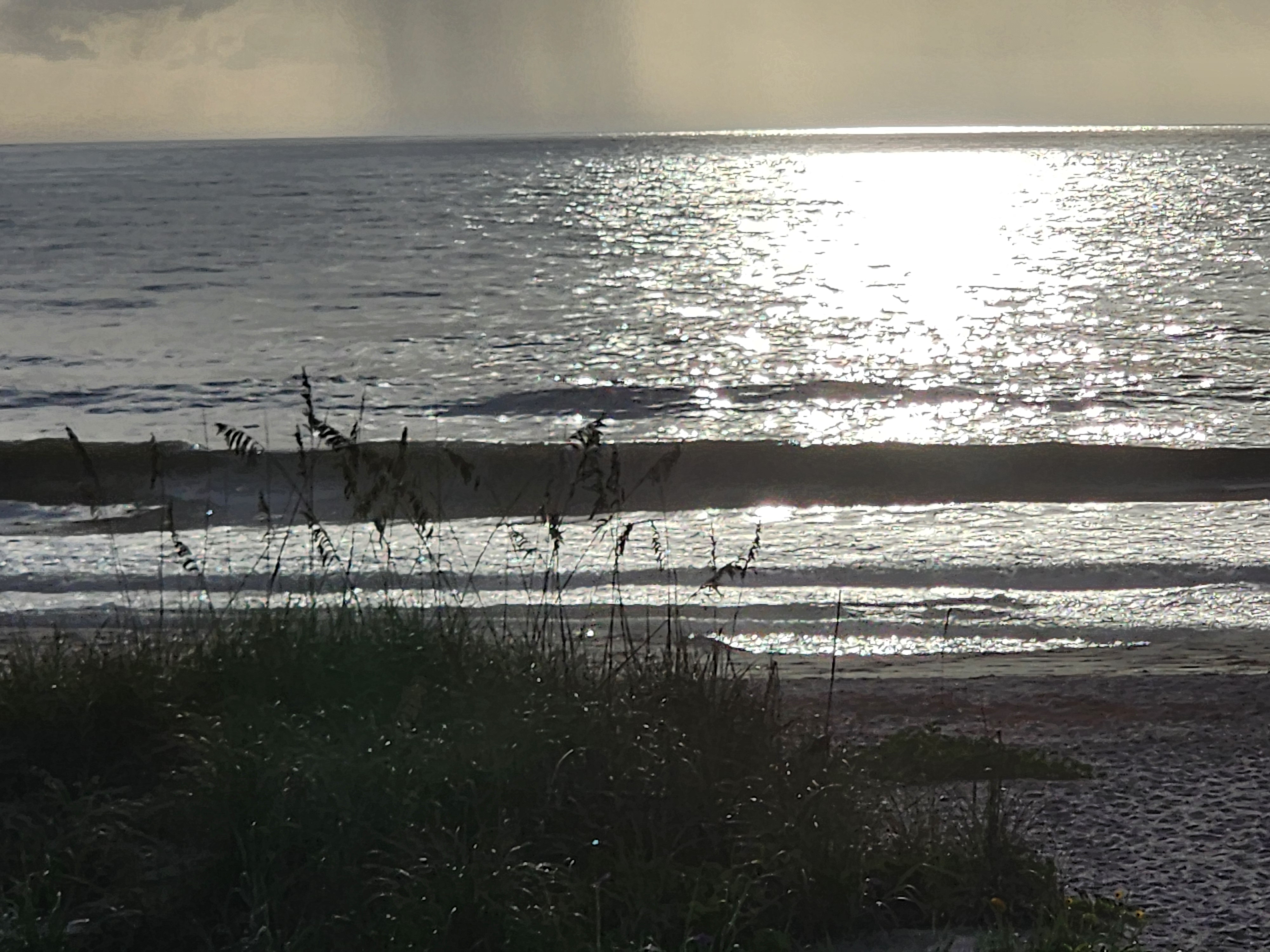 Silver morning sun glare across the water at 32nd Street with lines of surf silhouetted against the shimmer and a dark cloud bank offshore