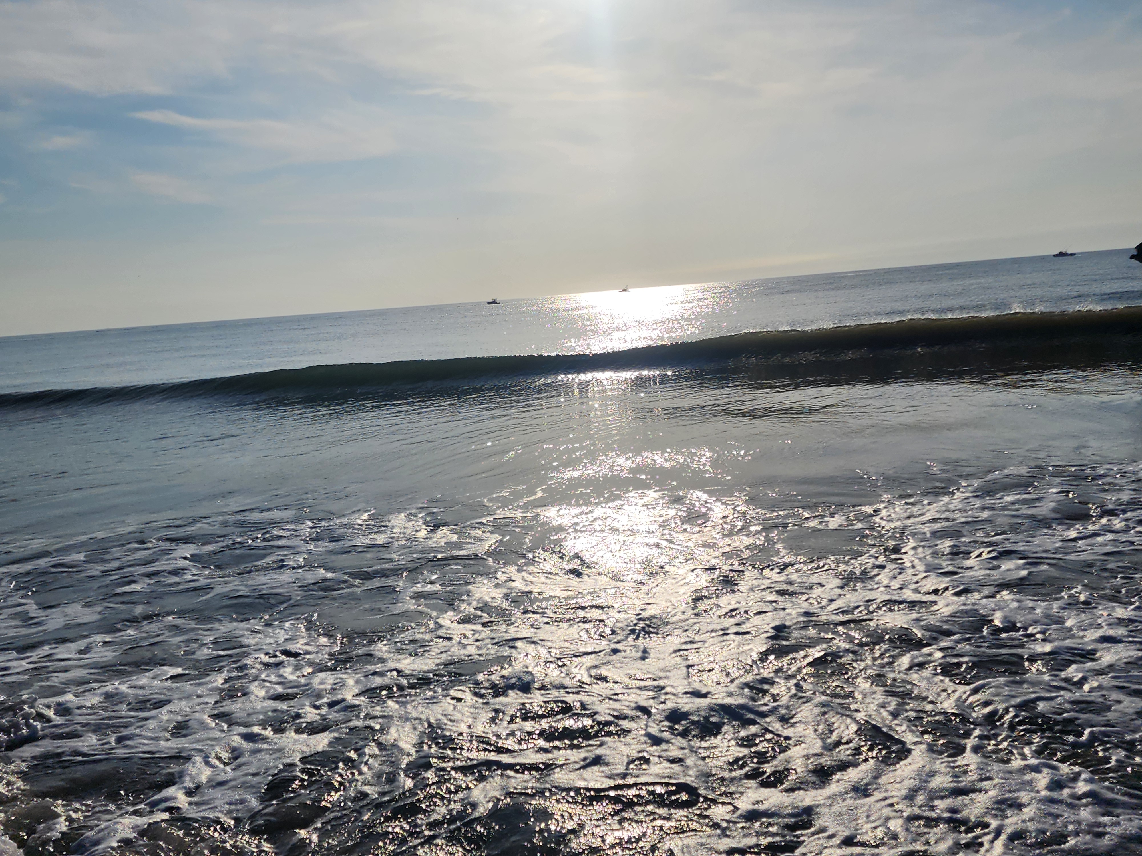 Small clean shorebreak wave feathering into the sand with sun glitter on the water and boats sitting offshore at 32nd Street