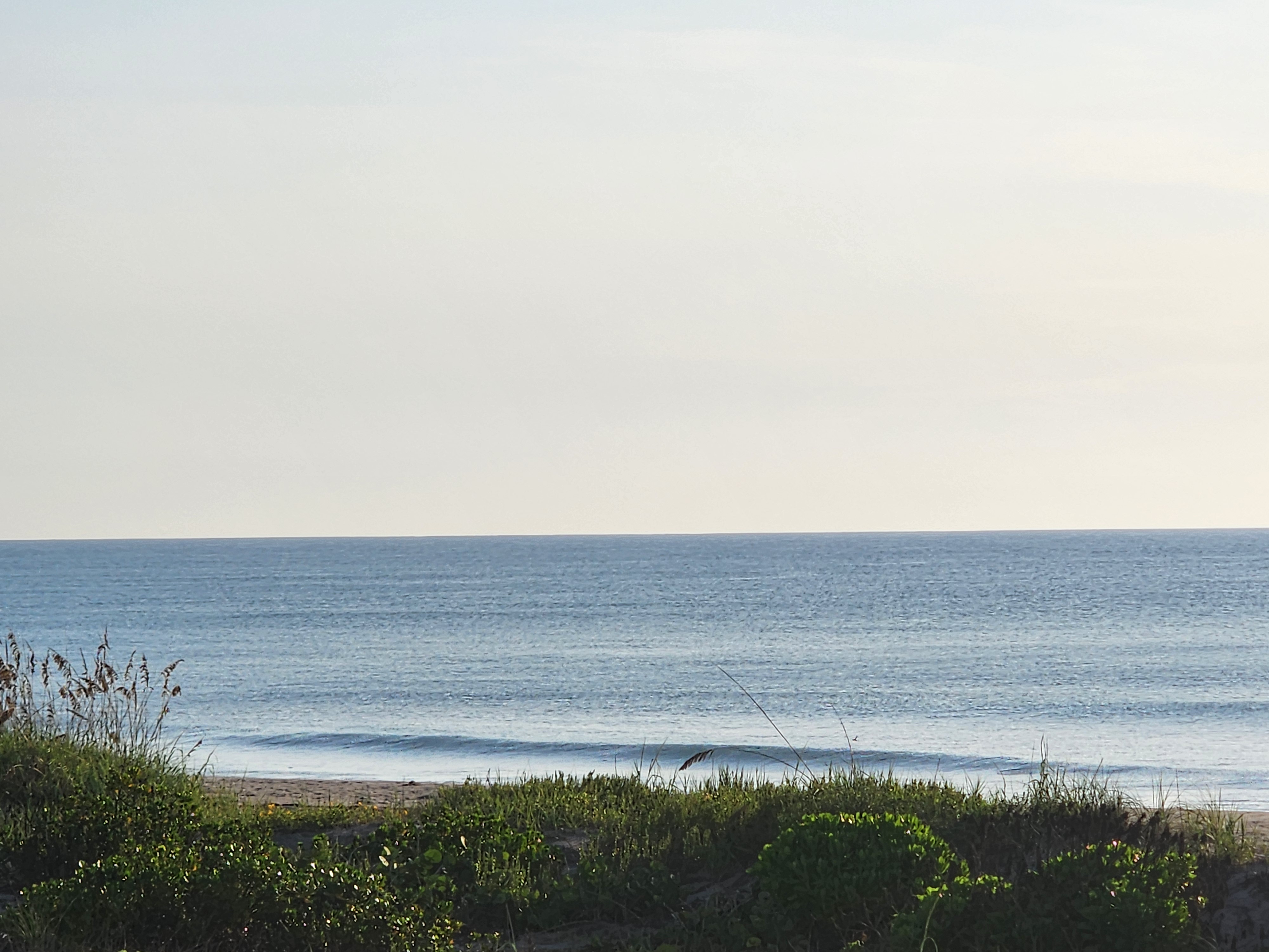 Wide view of a nearly flat blue Atlantic from the dune line at 31st Street, one small crumbling line pushing onto the sand