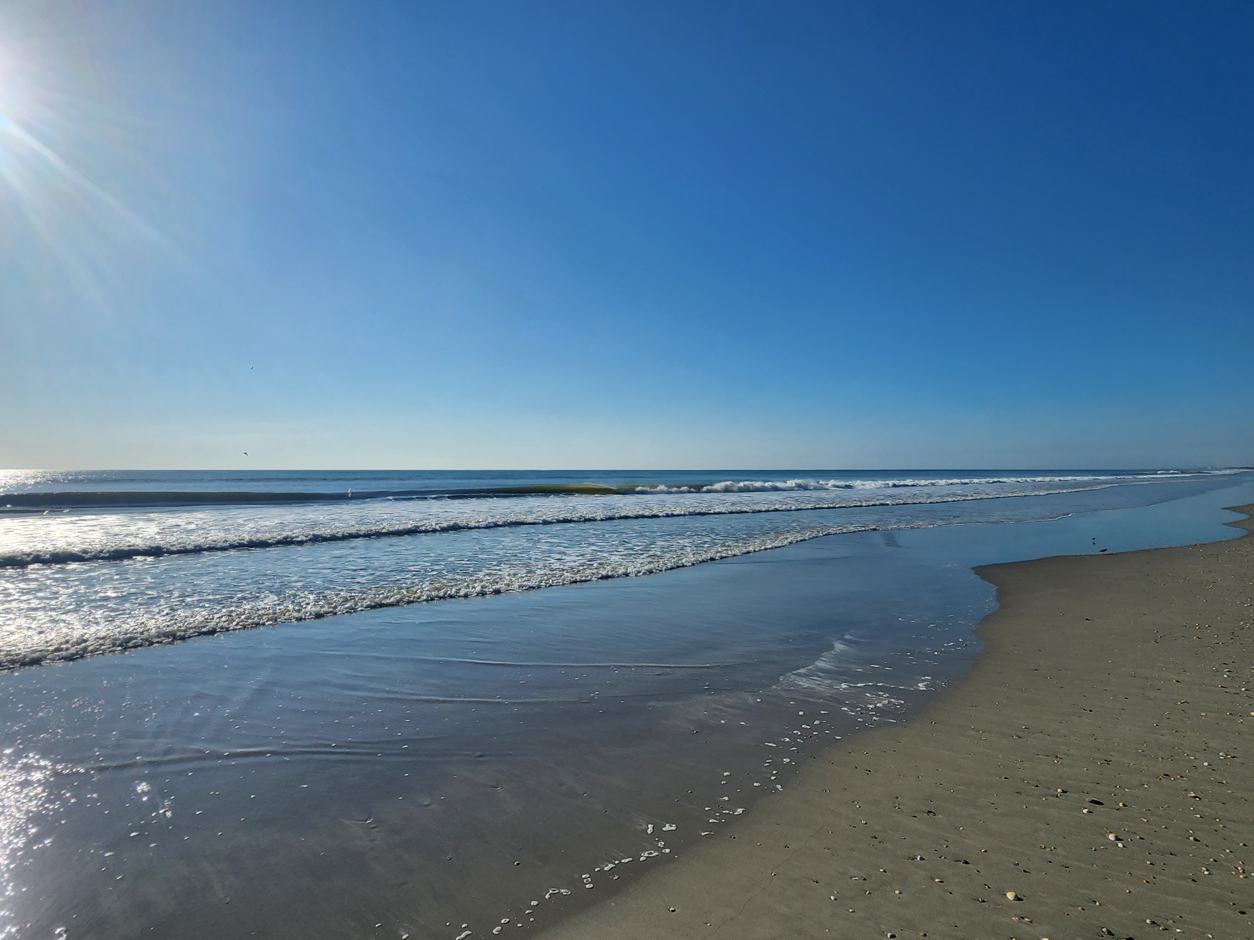 Wide view down the wet sand at low tide with long lines of whitewater rolling in under a deep blue sky, South Cocoa Beach, August 2026
