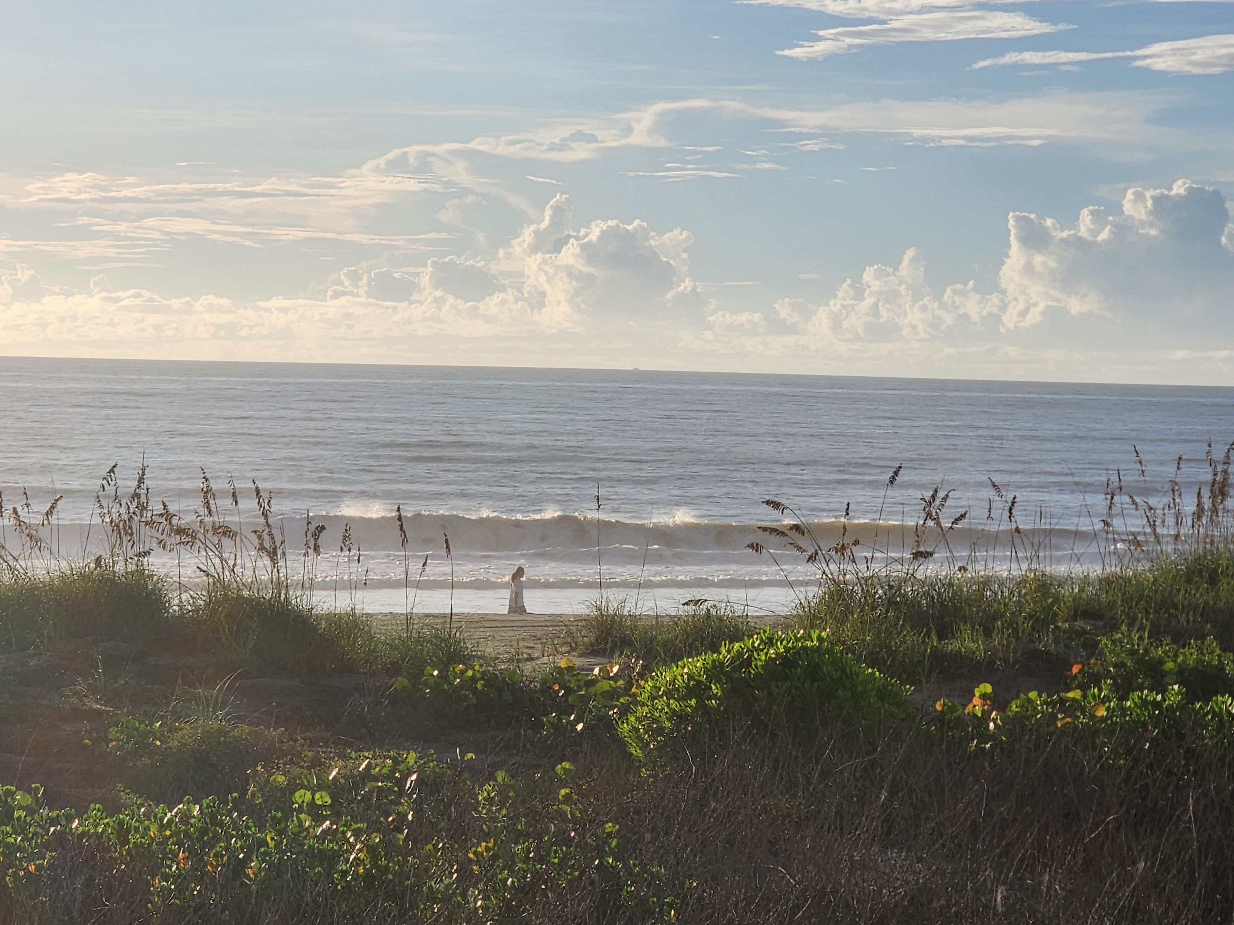 A beachgoer in white standing at the water's edge as a 2 foot wave breaks behind her, South Cocoa Beach