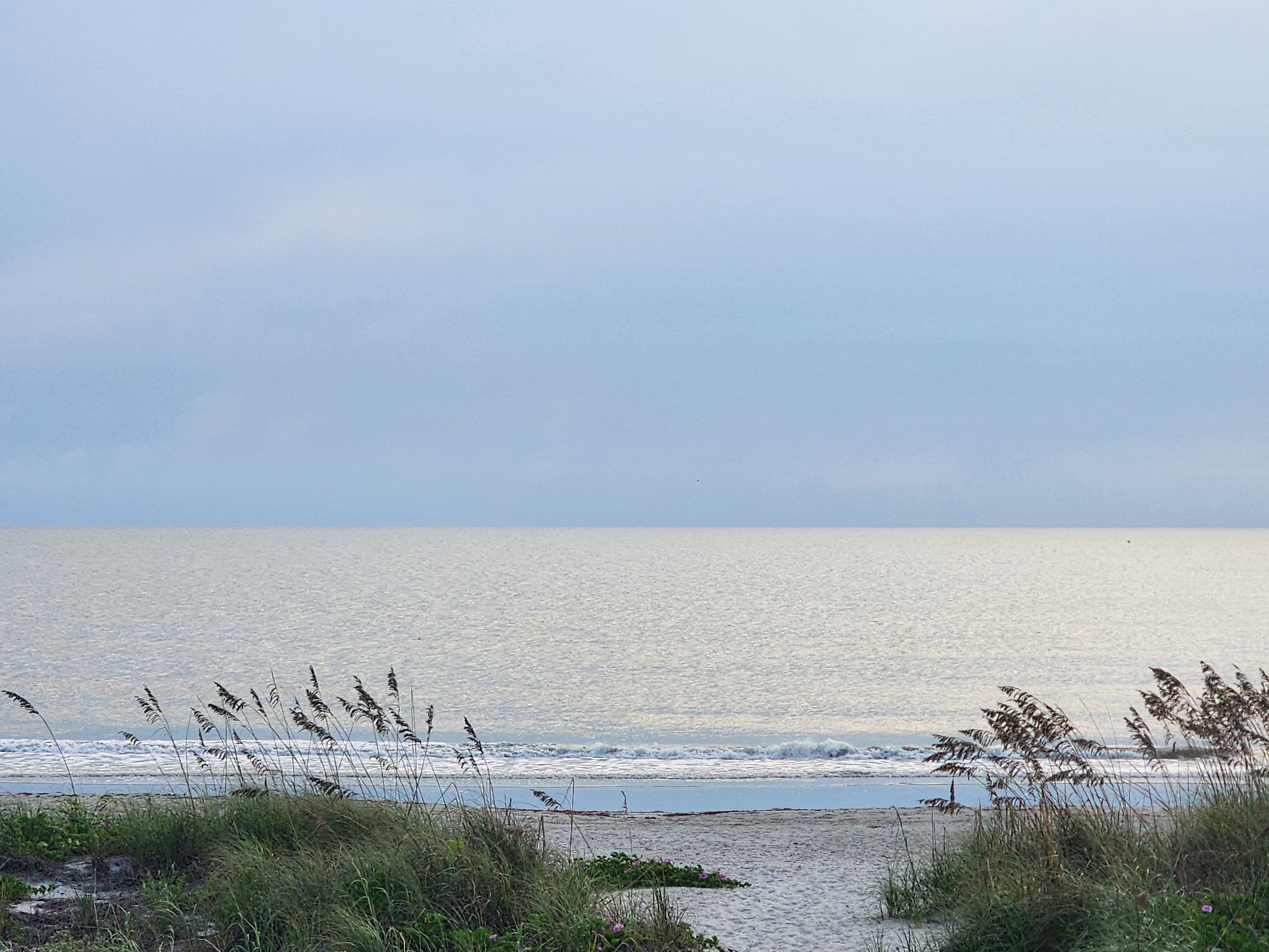 Glassy sheen across a windswept flat sea with a thin band of whitewater and a dune path of sea oats in the foreground at 32nd Street, South Cocoa Beach, September 2026