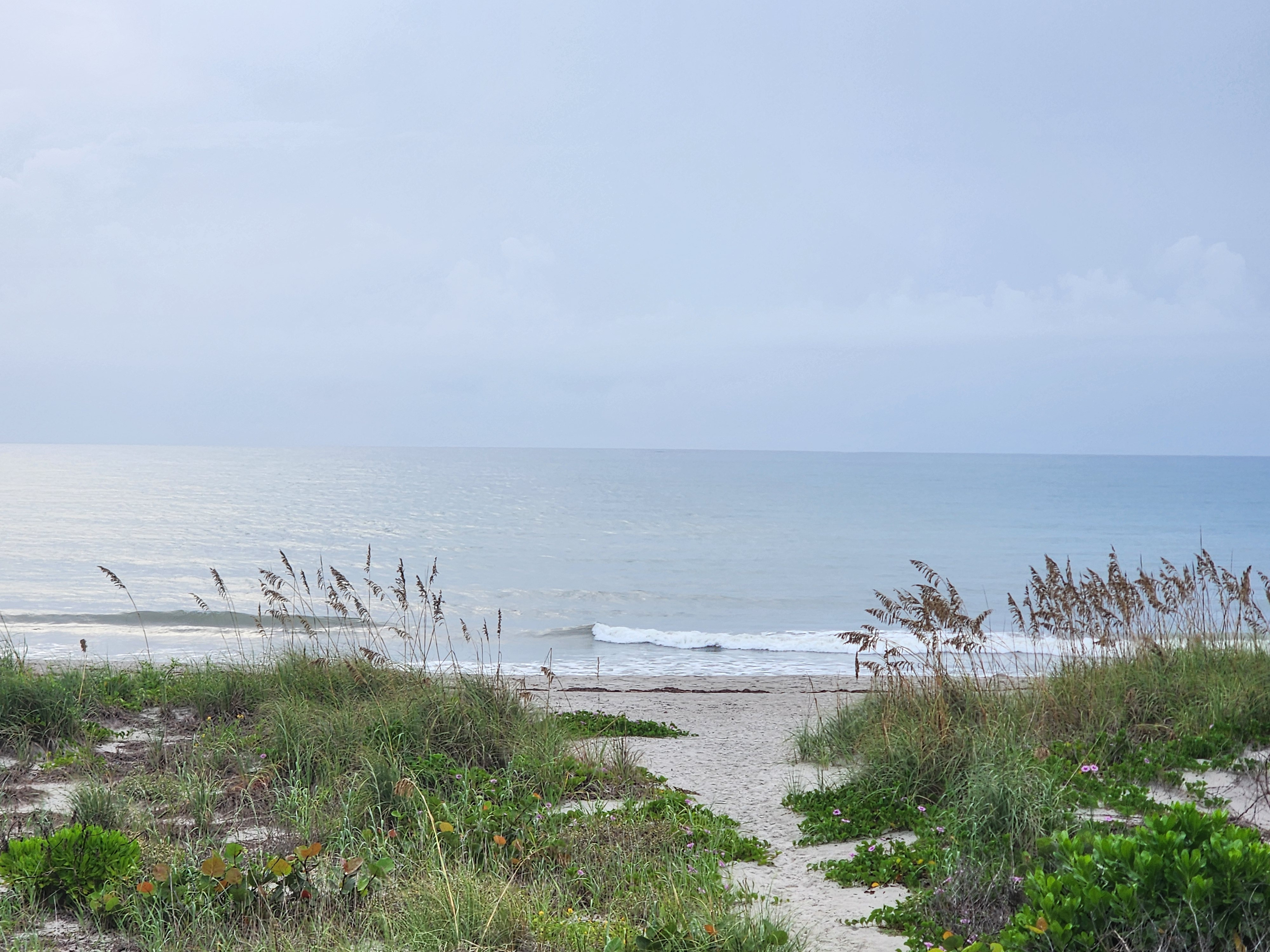Sandy crossover path through the dune out to the beach, small waves breaking beyond, hazy high clouds