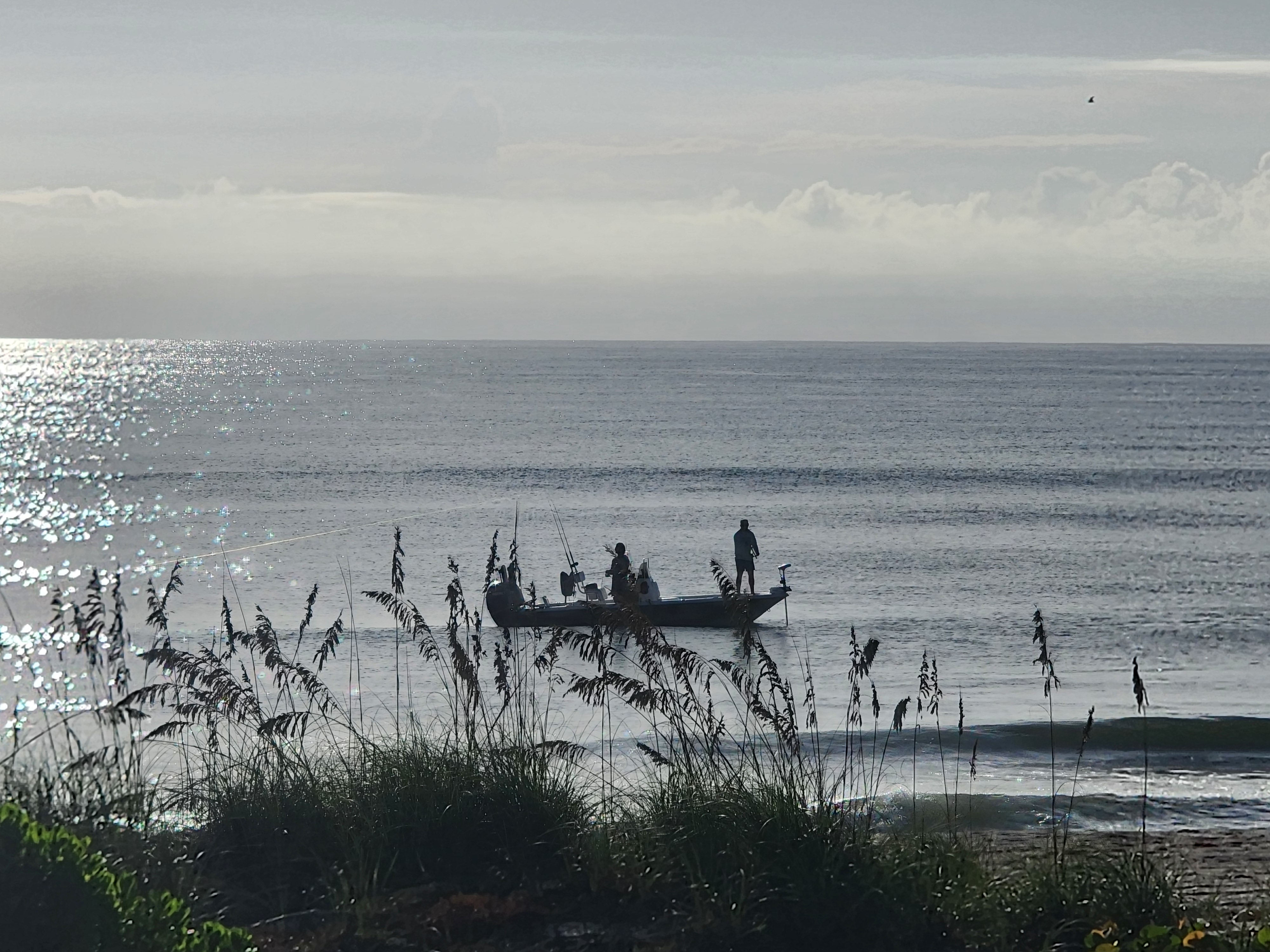 Two anglers working a center console boat just outside the breakers on a silver morning sea, sea oats in the foreground at 32nd Street, South Cocoa Beach, September 2026