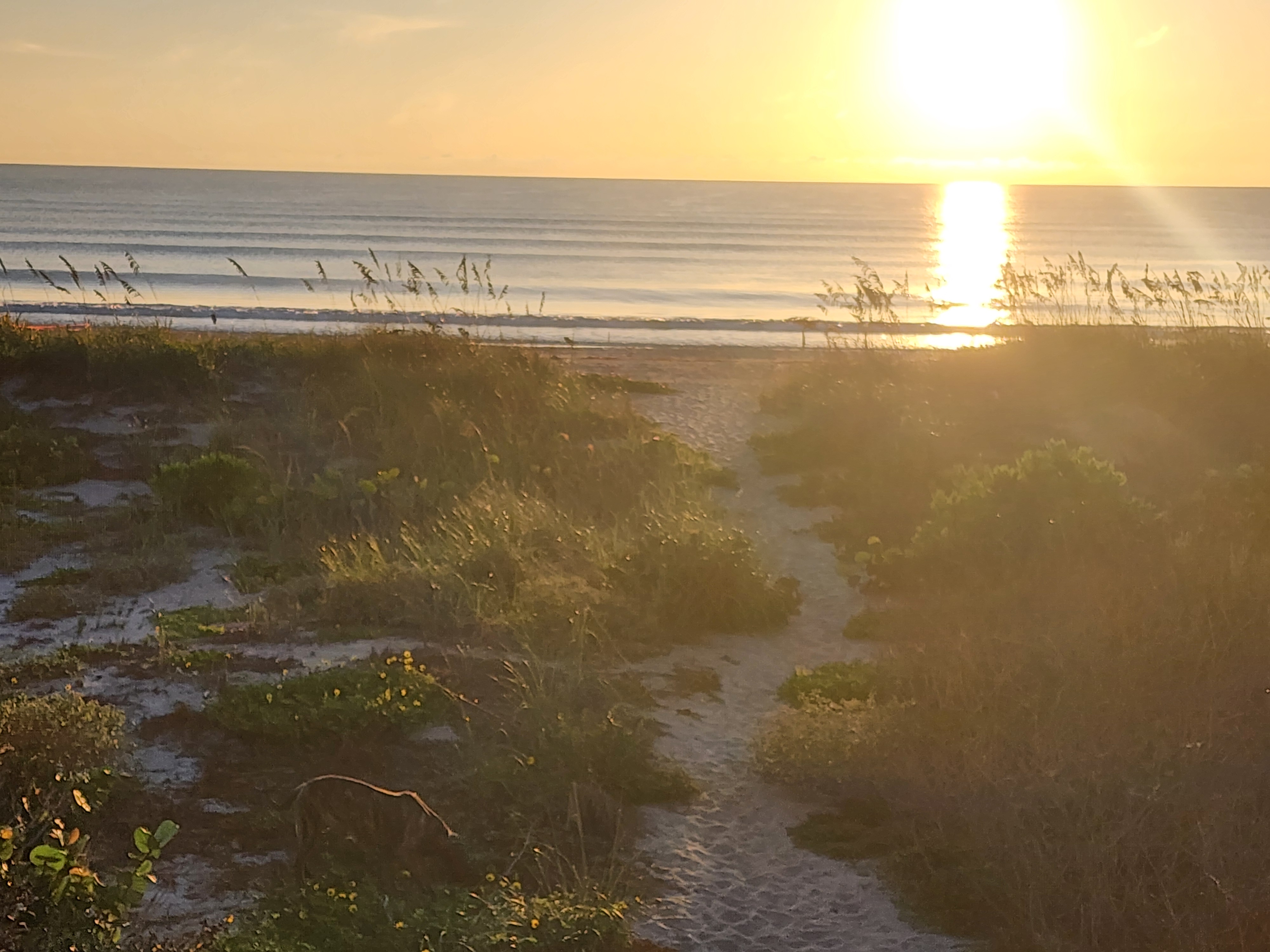 Sandy dune crossover trail running down to the beach with stacked swell lines and the rising sun glinting off the water, 32nd Street, South Cocoa Beach, September 2026