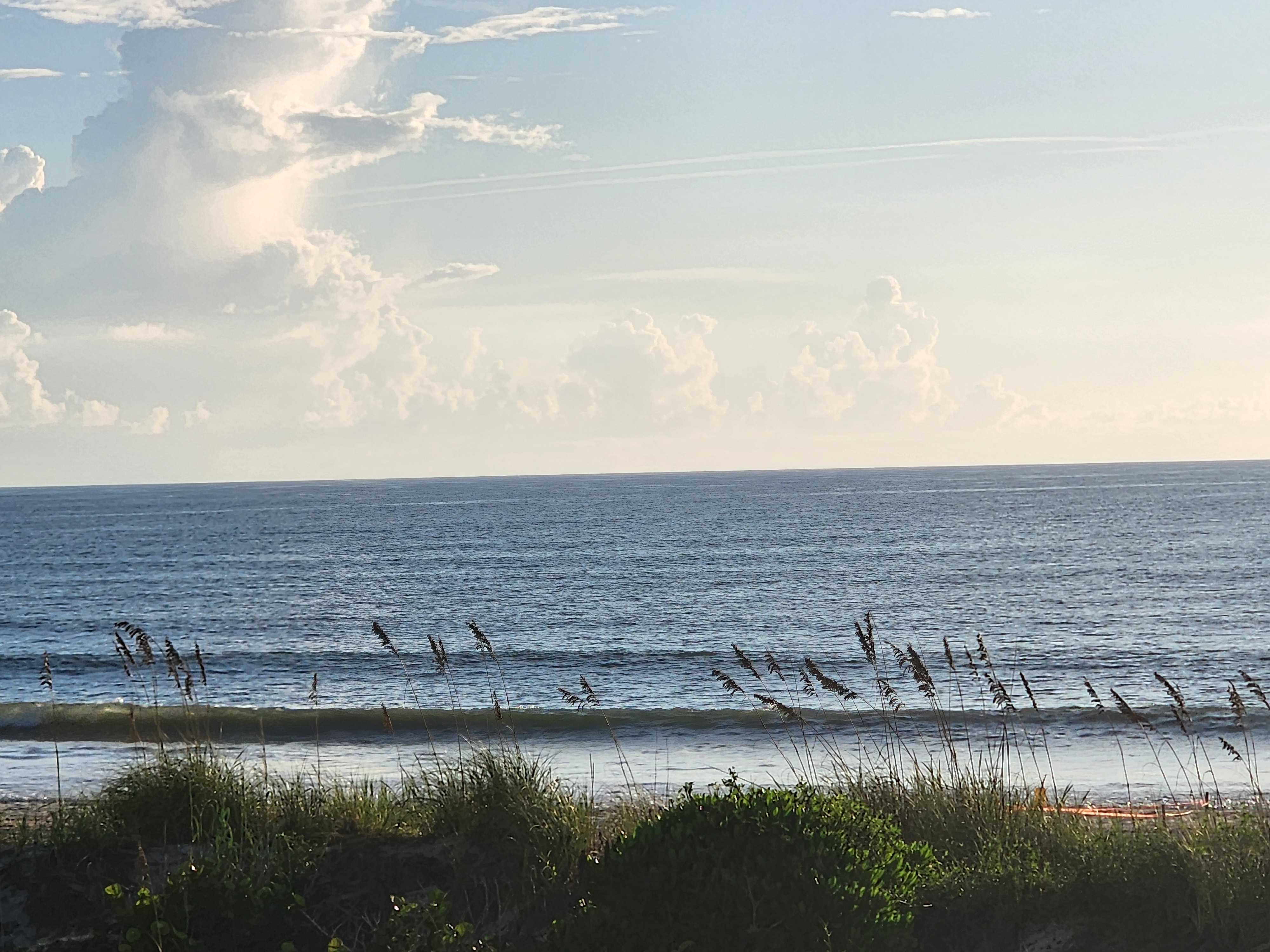 Small lines stacking up under towering cumulus clouds and contrails, sea oats silhouetted along the dune, South Cocoa Beach