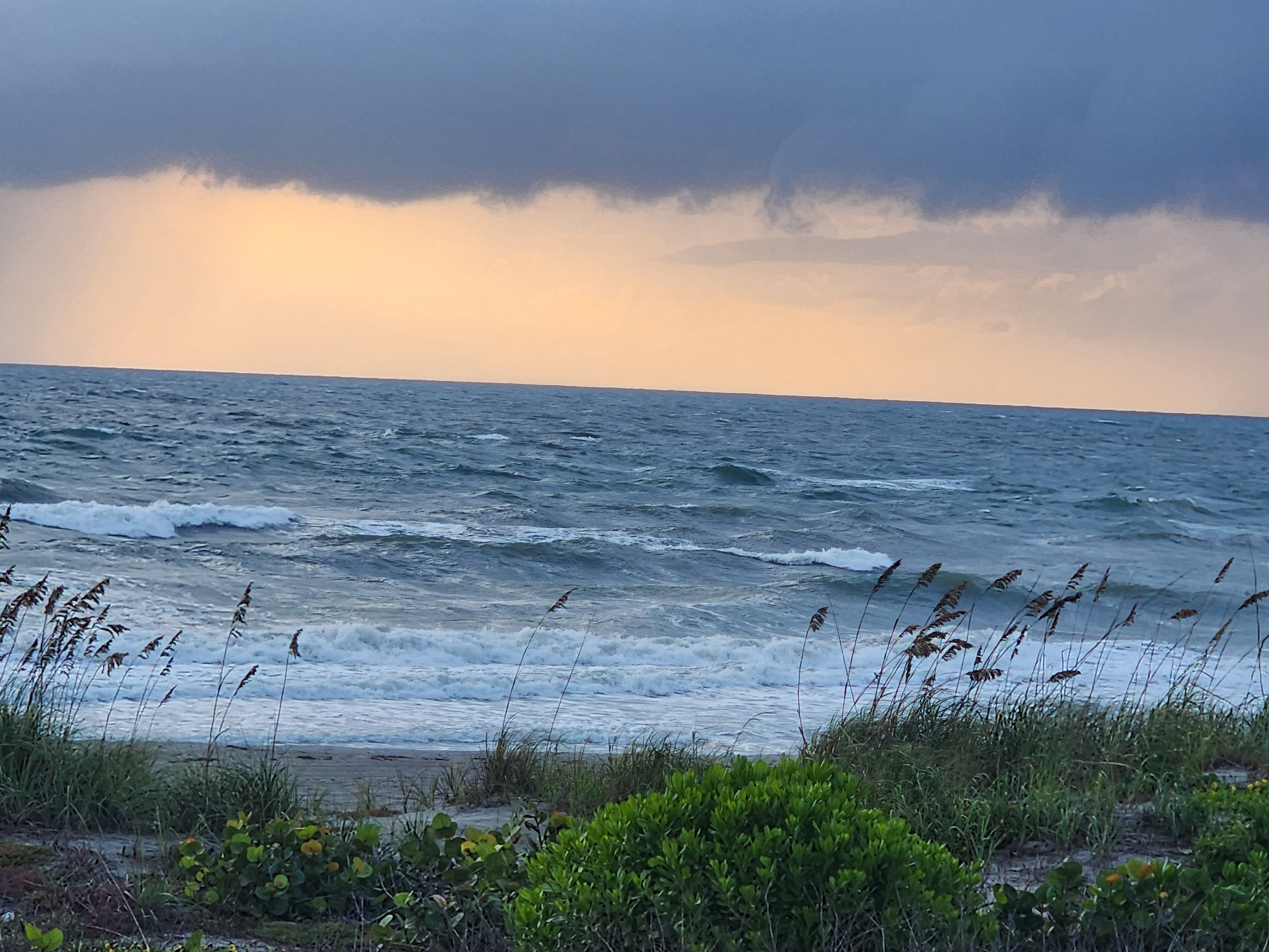 Dark rain band over a glowing orange horizon above windswept surf, sea oats and sea grapes bending in 20 knot ENE winds at Cocoa Beach