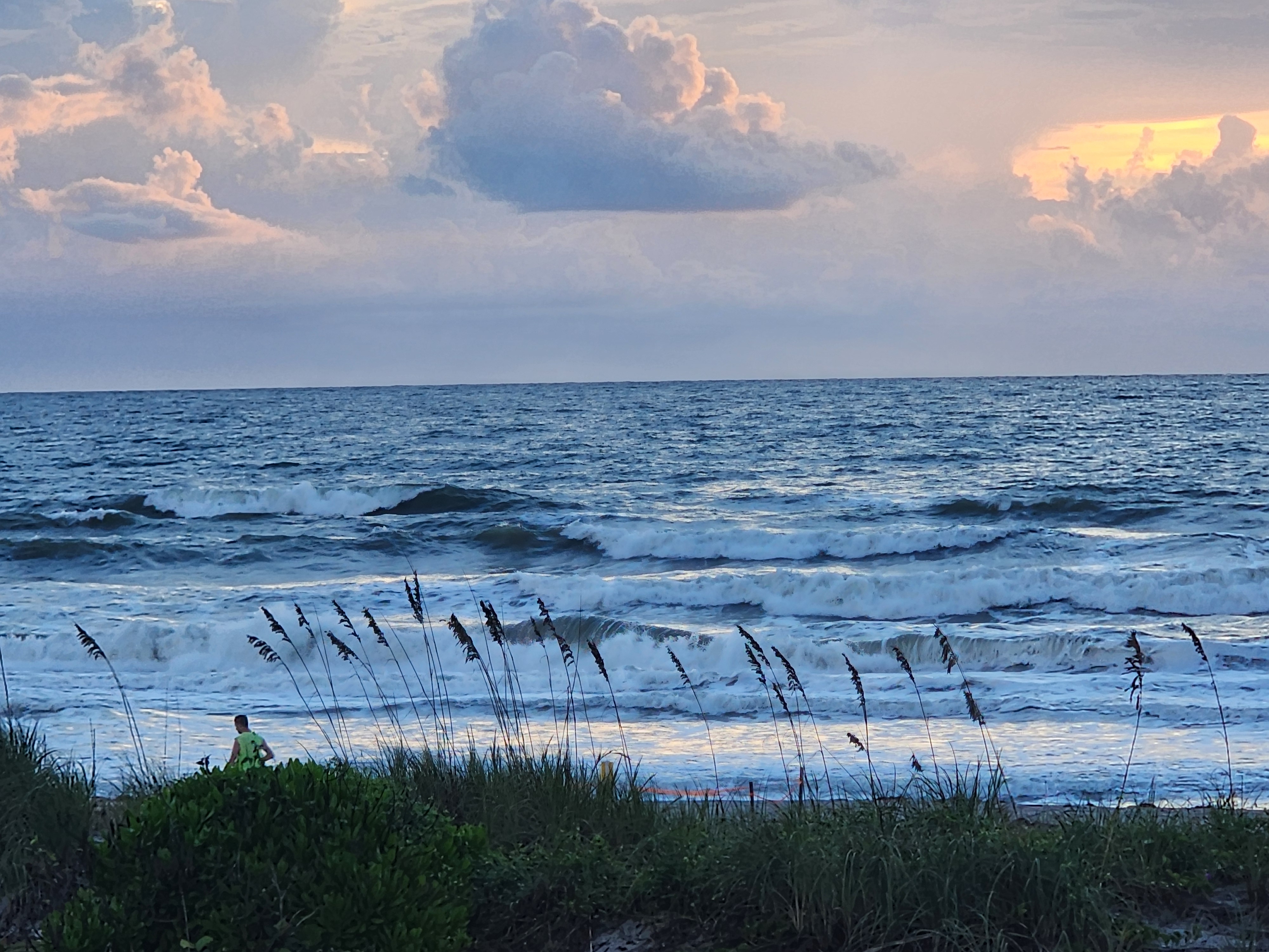 Three to four foot waves stacking up and breaking outside on the low incoming tide, pink sunrise clouds overhead and a walker in a neon shirt on the sand at Picnic Tables, South Cocoa Beach