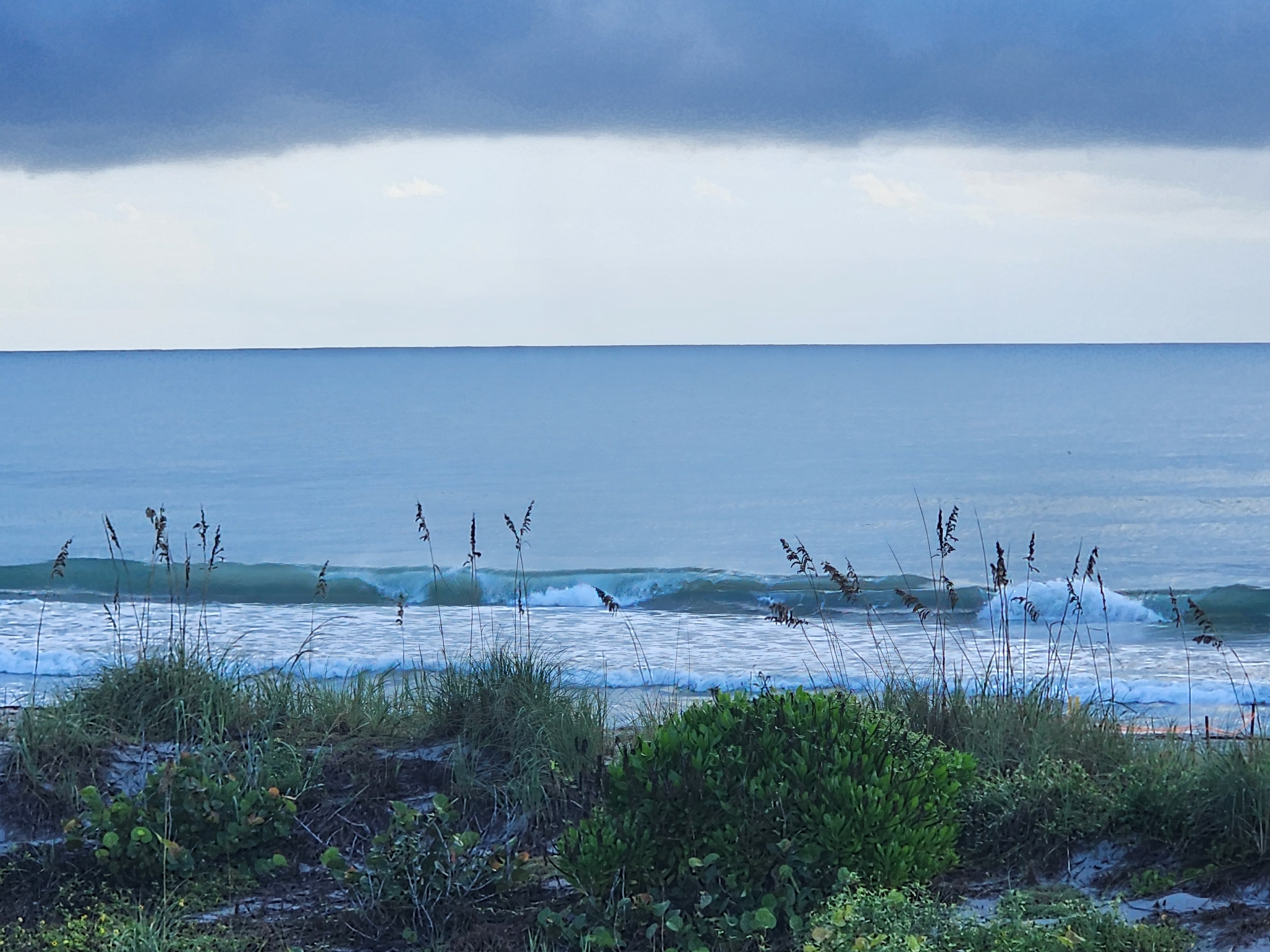 Clean 1-2 ft waves peeling nearshore at low tide in Cocoa Beach, framed by sea oats and dune vegetation under a dark storm cloud bank