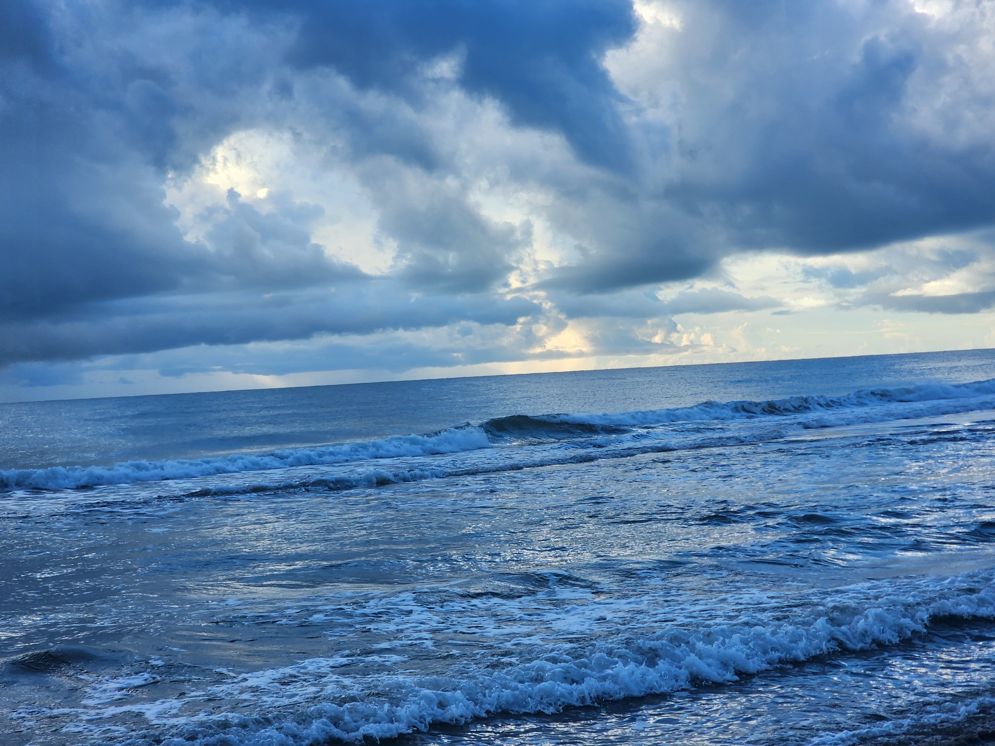 Small clean lines rolling into the Cocoa Beach shorebreak at low tide with dramatic storm clouds and breaks of morning light offshore