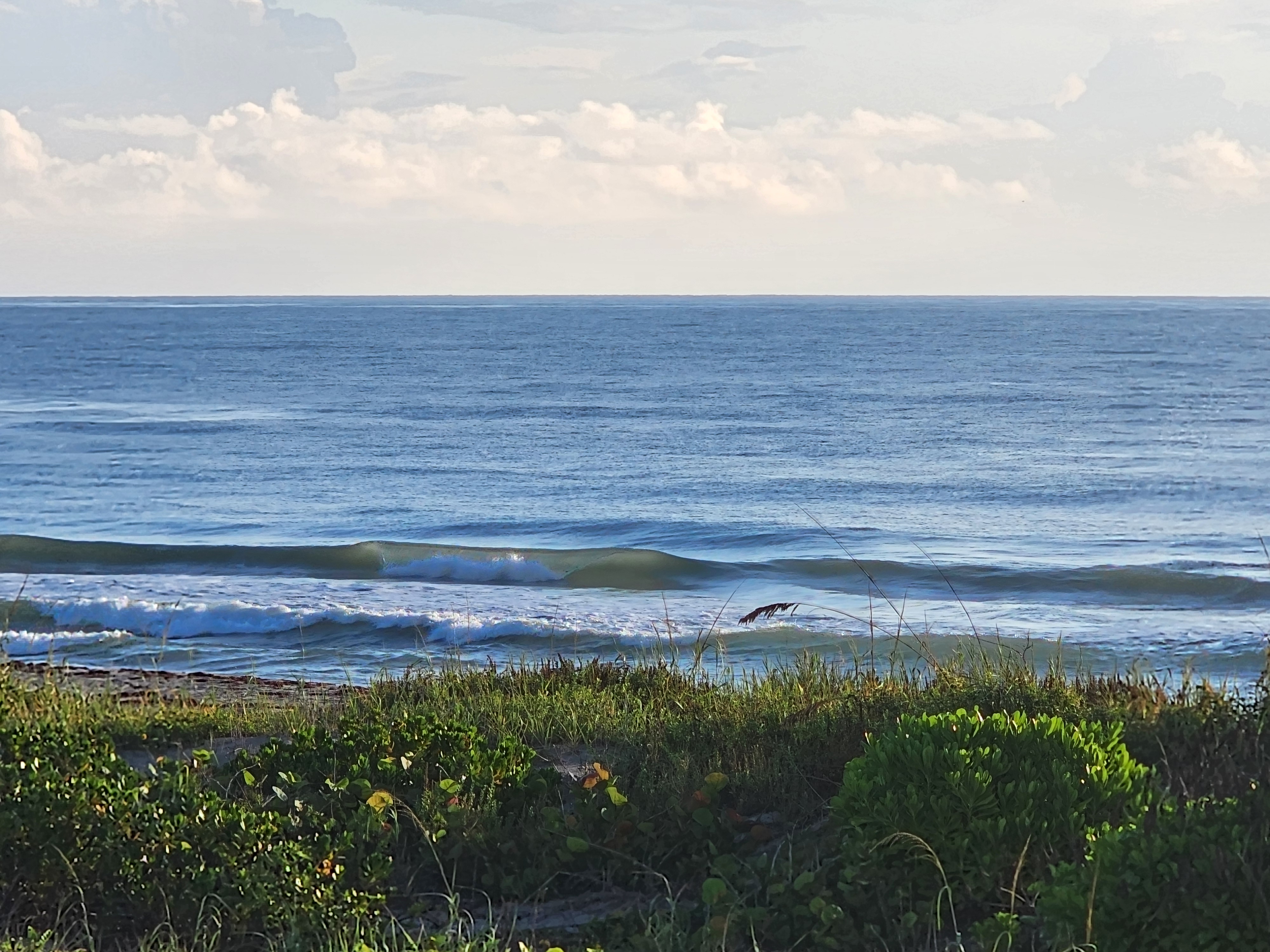 Clean little wave pitching on the inside with whitewater rolling toward shore under puffy clouds