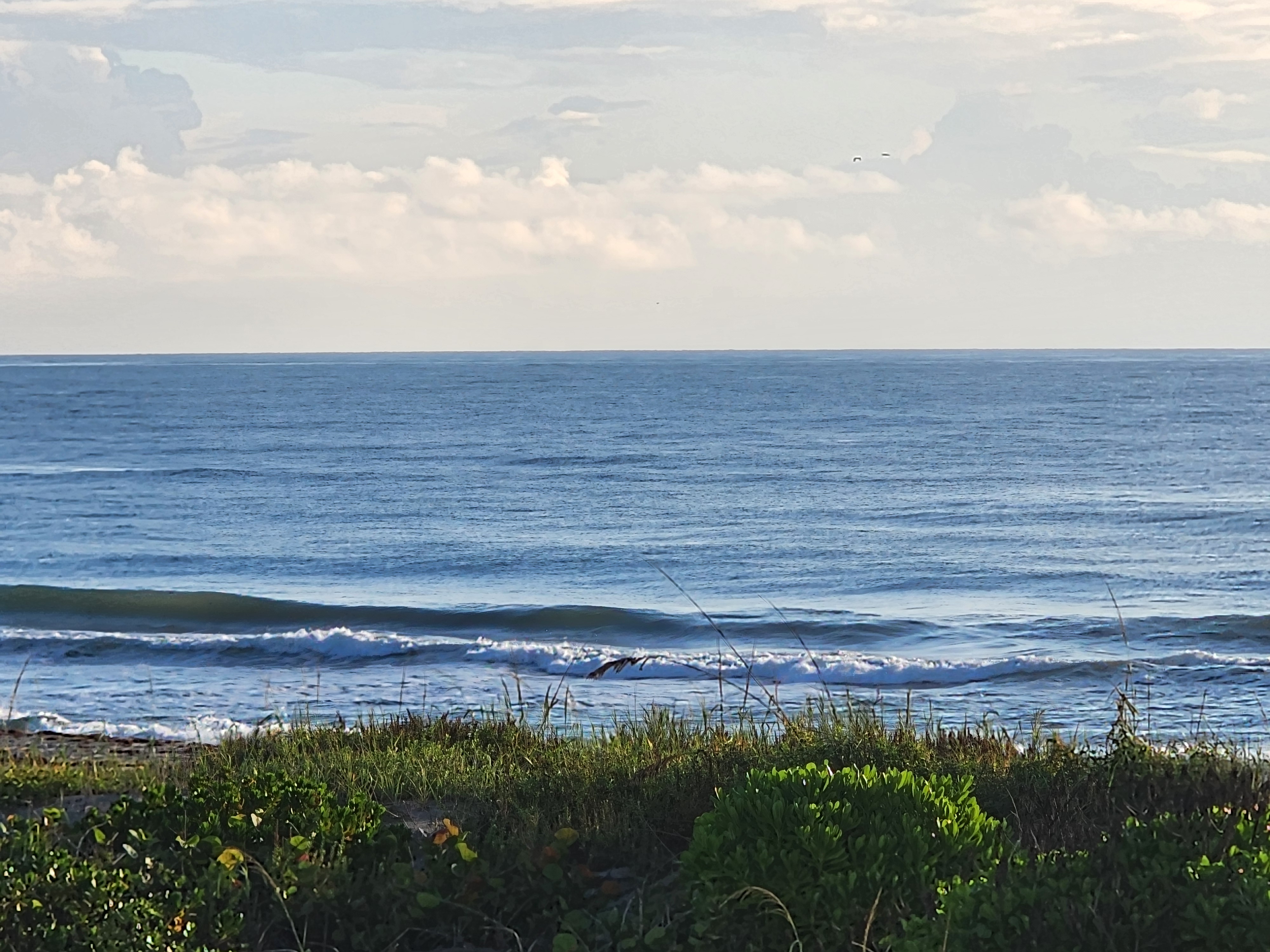 Wide view of small lined-up sets and calm blue ocean beneath scattered morning clouds