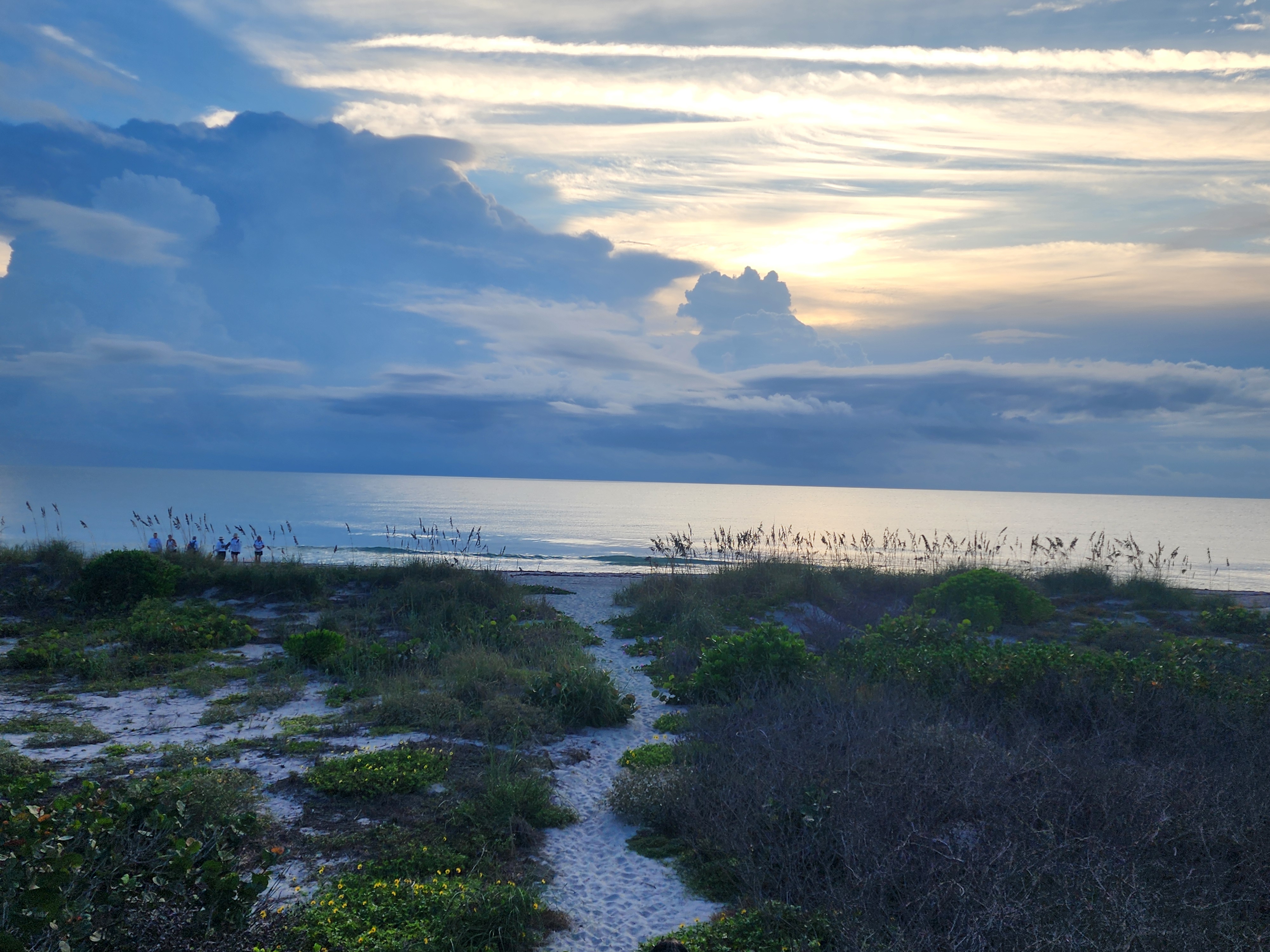 Sandy path through the dunes leading to the beach, with a few people at the water's edge and towering clouds catching the morning light