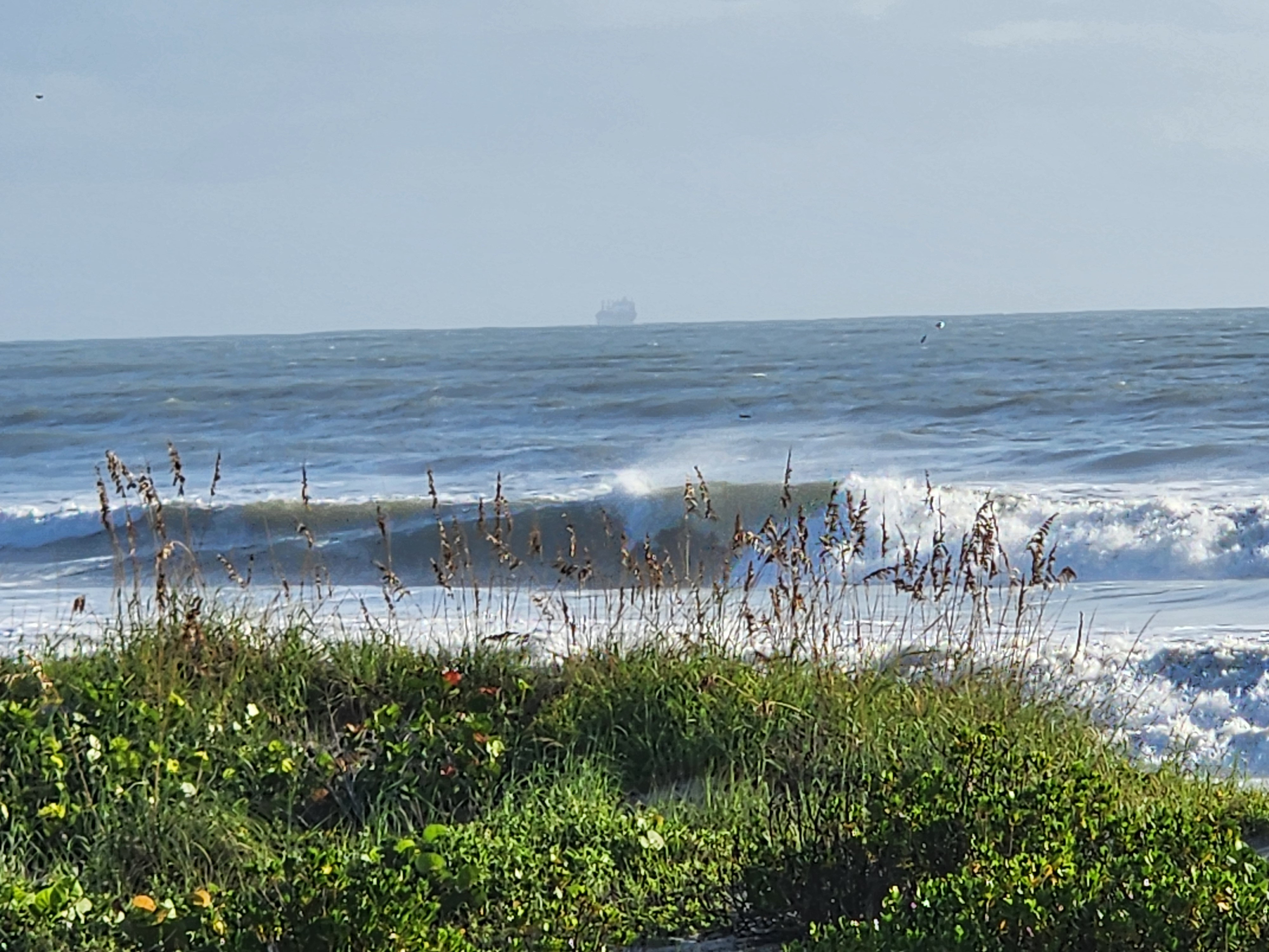 Overhead Cocoa Beach wave line feathering behind golden sea oats, with a cargo ship on the hazy horizon