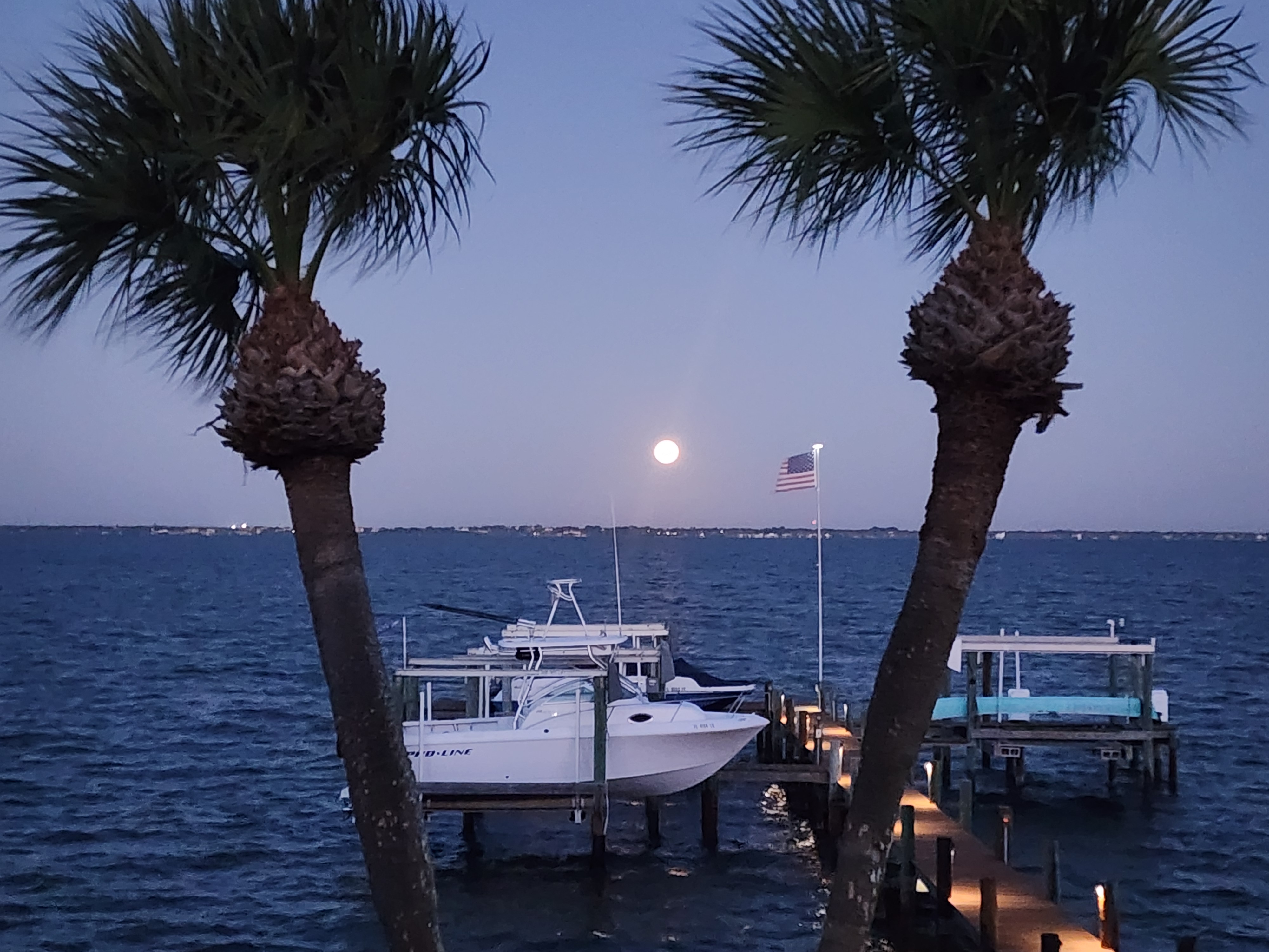Full harvest moon rising over the Banana River at dusk, framed by two sabal palms, with a lit wooden dock, boat lifts, a center console on its lift, and an American flag flying off the end of the pier.