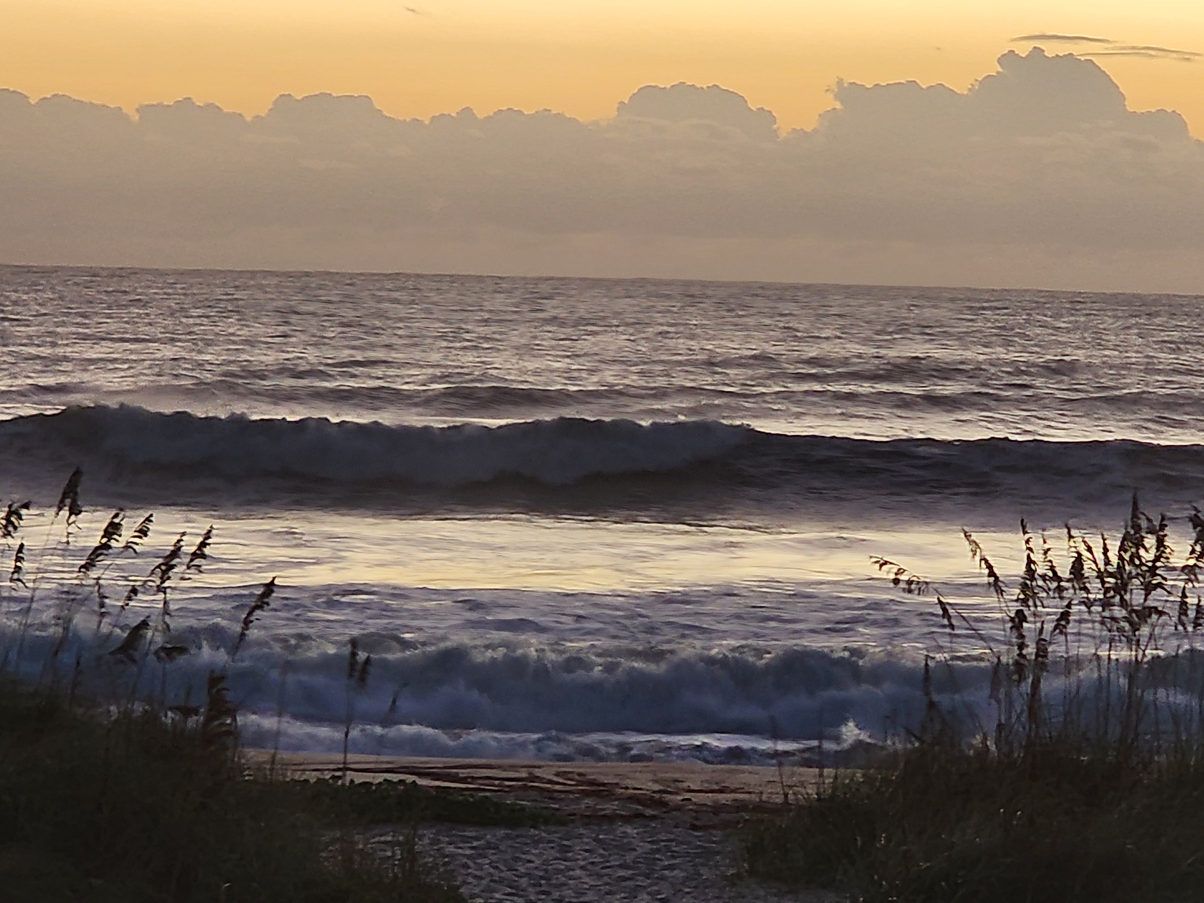 Stacked lines of wind-chopped swell breaking across the outer and inner bars, shorebreak dumping on the sand between sea oats under towering orange-lit clouds.