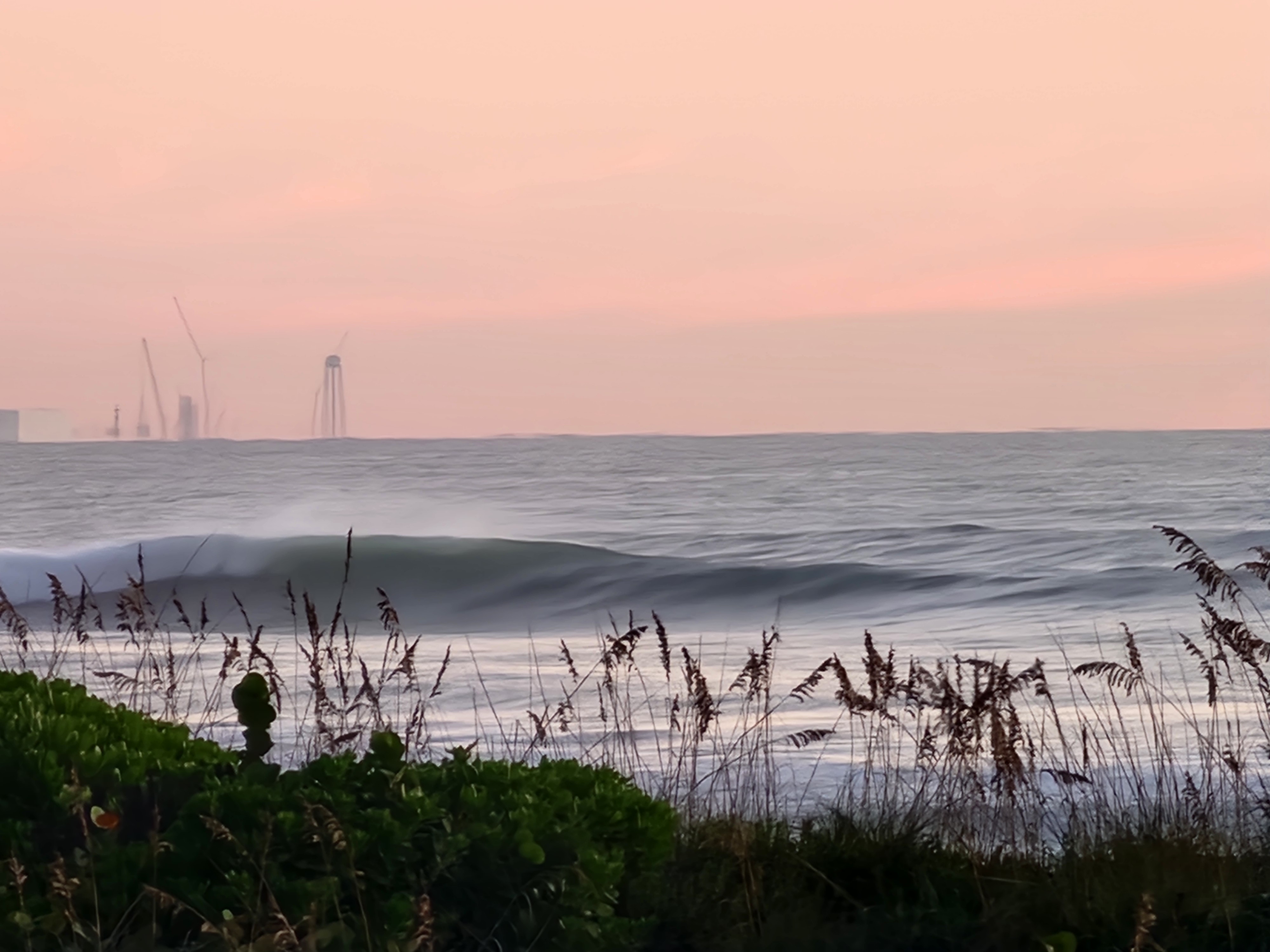 A clean wave feathering in the light NW offshore wind behind sea oats and sea grapes, with the launch towers and cranes of Cape Canaveral rising on the pink northern horizon.