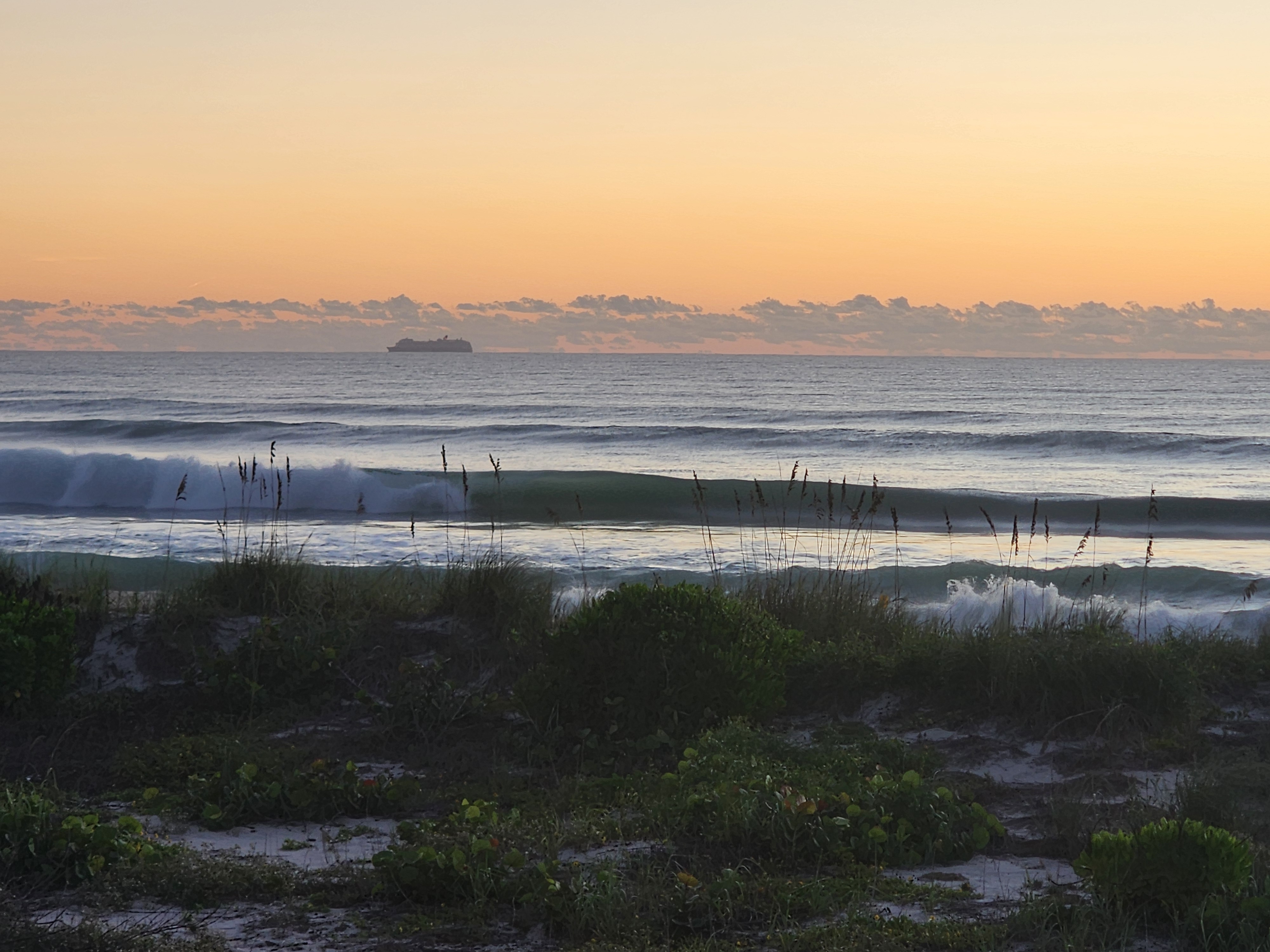 A long green wall closing out across the inside sandbar with shorebreak exploding on the sand, sea oats on the dune and a cruise ship on the horizon under a low line of orange-lit clouds.