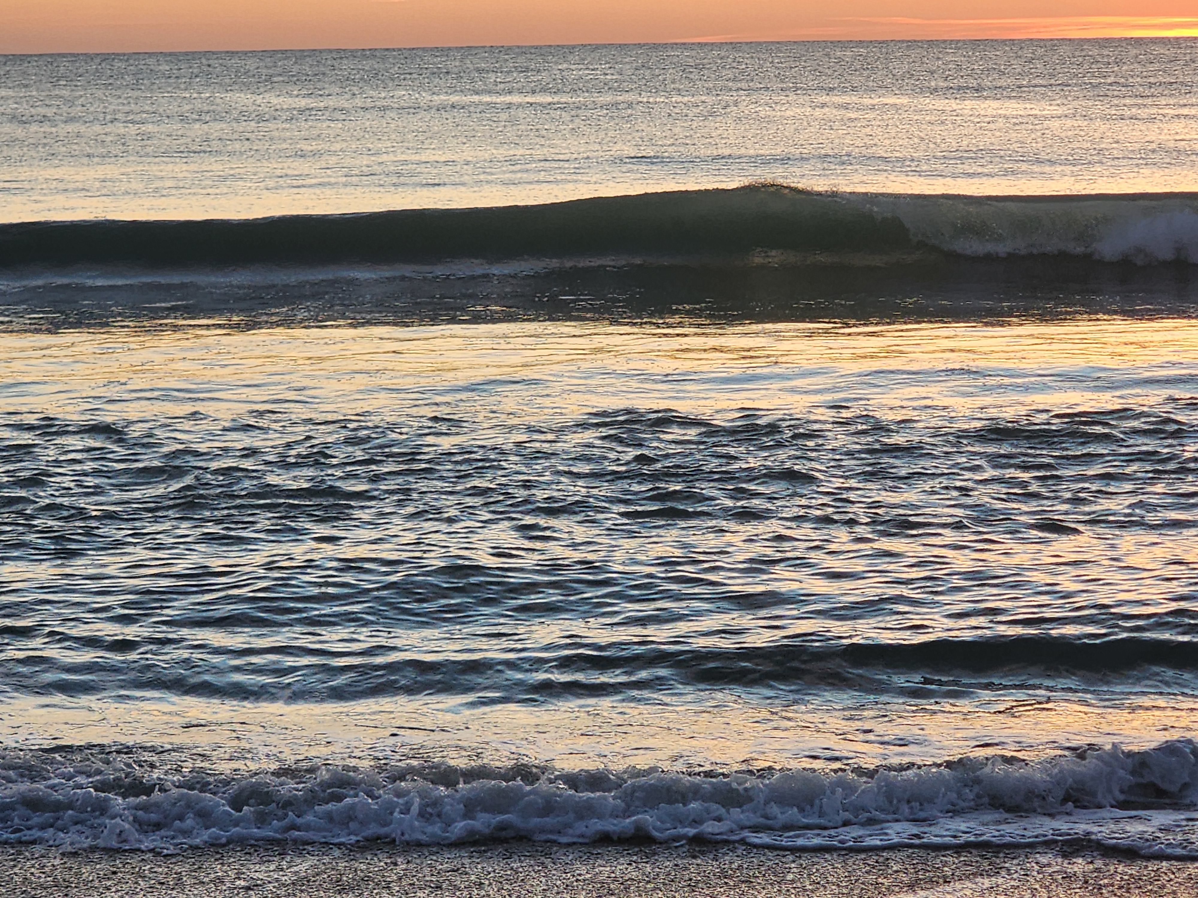 A clean green lip feathering in the light NW offshore wind as it throws over on the inside, glassy water shimmering gold in the foreground and shorebreak washing onto the sand.