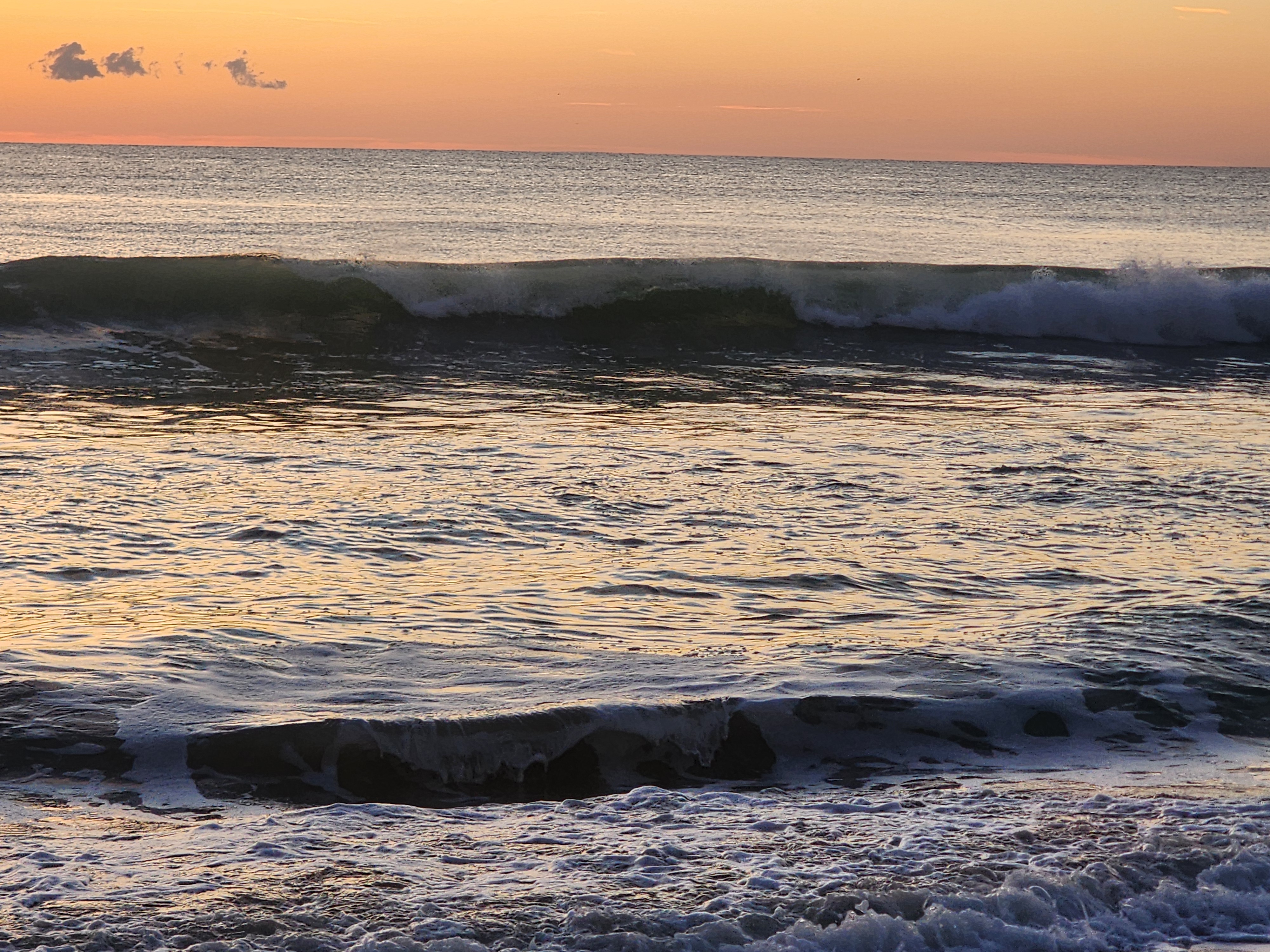 A long-period line pitching and spraying across the inside bar with foamy backwash in the foreground, the Atlantic glassy and a few small clouds drifting in an orange sky.