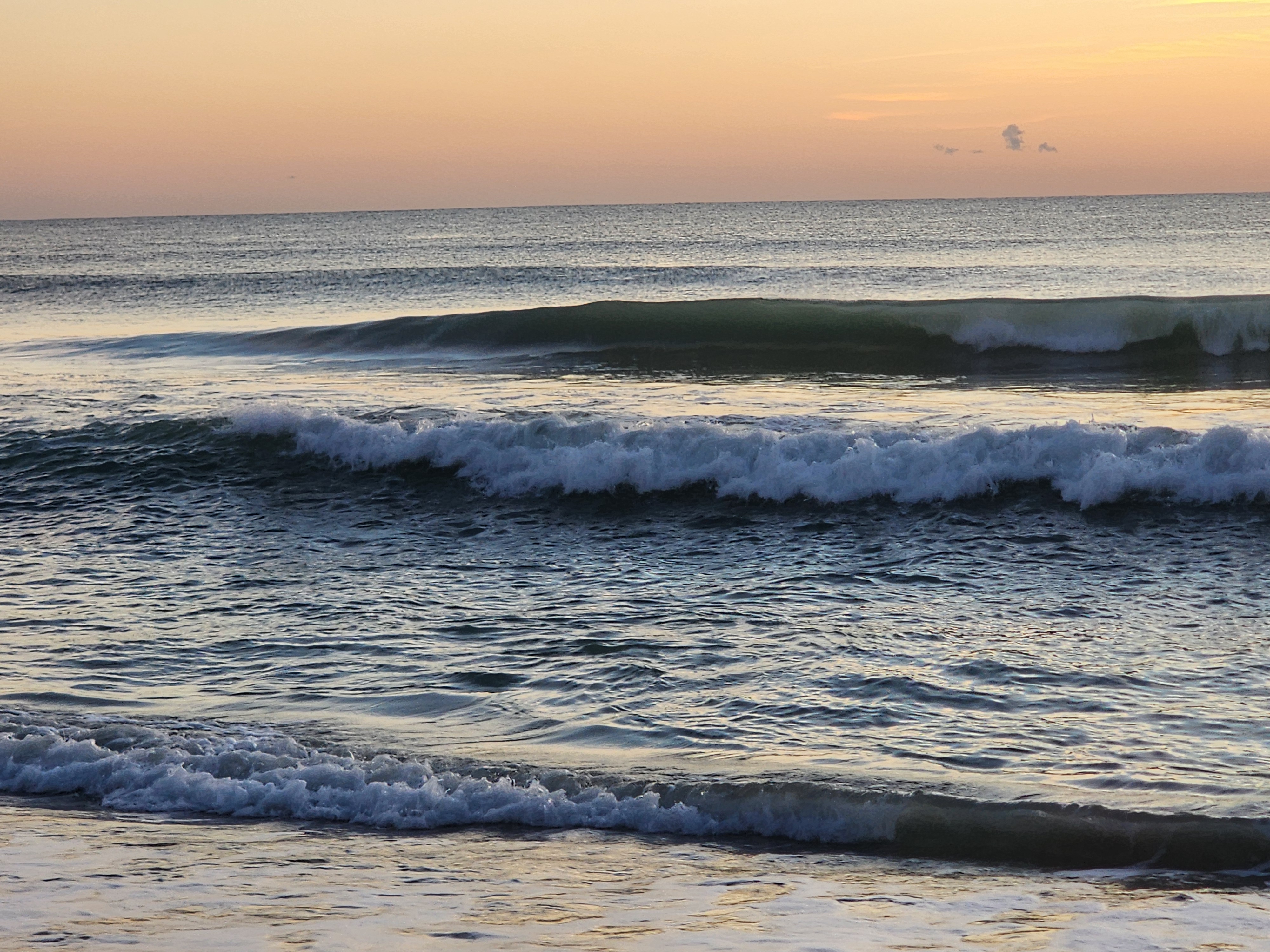 Two lines of long-period swell stacking up on the inside, the outer wave standing up green and clean while the inner line breaks white under a pale peach sky.