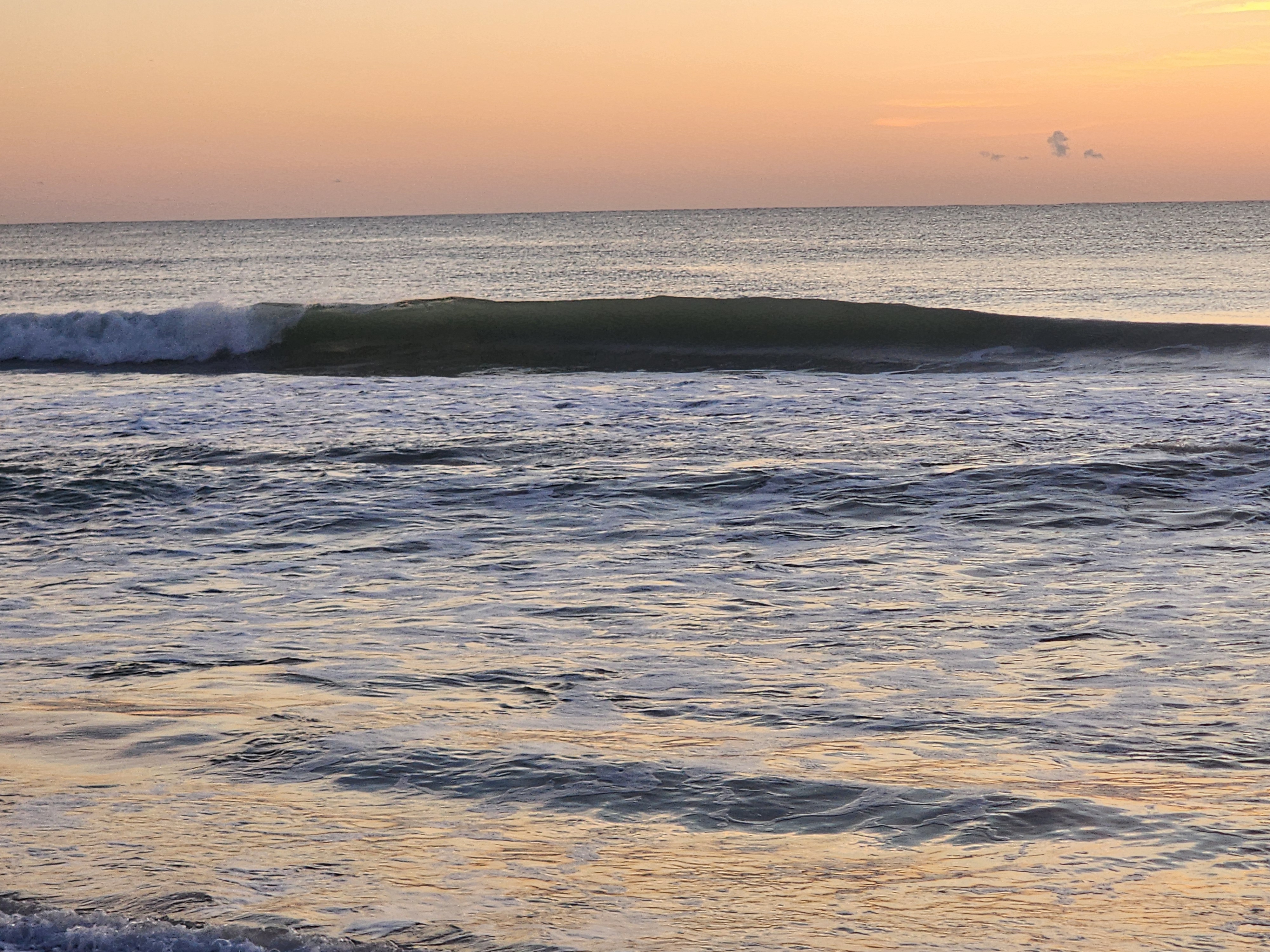 A smooth green wall peeling right to left on the inside at high tide, whitewater spilling from the shoulder and the ocean glassy out to a soft orange horizon.