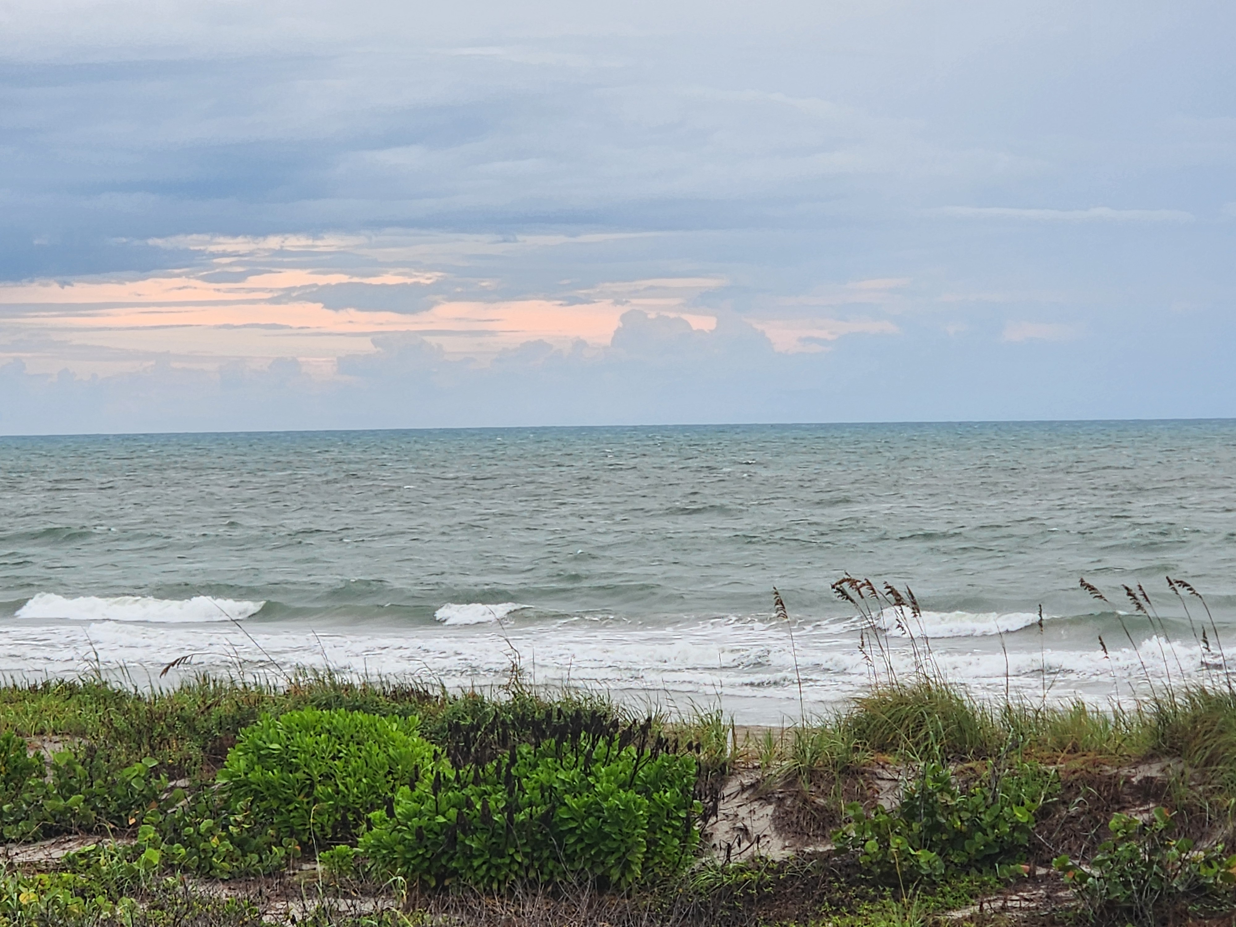 Choppy gray-green ocean with small breaking waves under heavy clouds, pink light glowing low on the eastern horizon